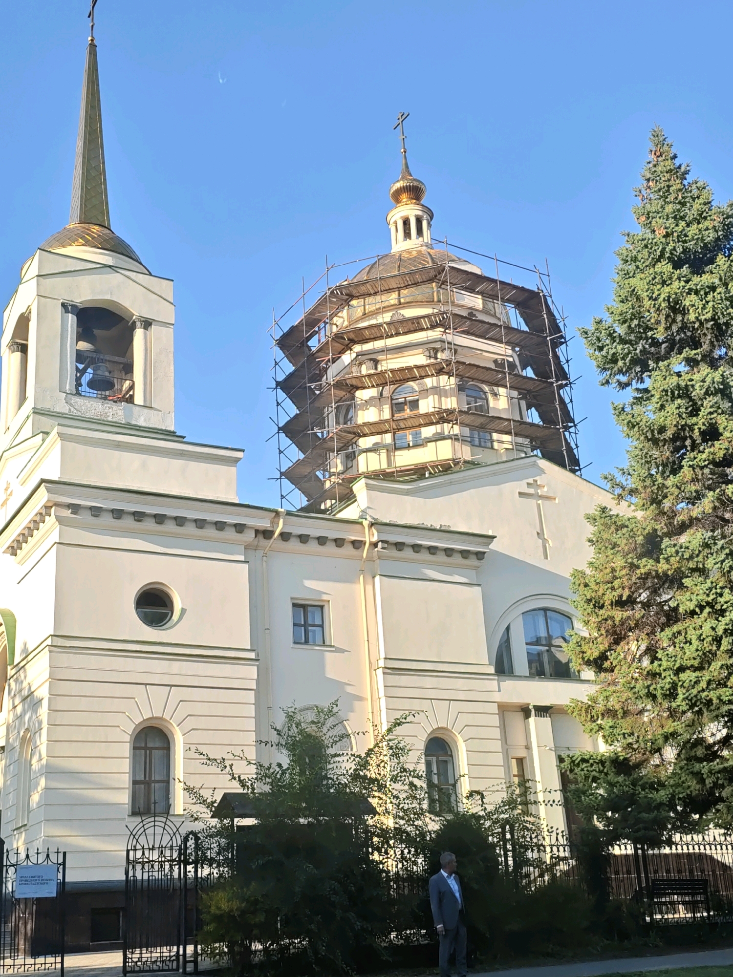 The image shows the exterior of the Ascension Cathedral in Rostov-on-Don, Russia, under renovation. Scaffolding surrounds the main dome, which is gilded and topped with a cross. To the left, a bell tower with a golden spire and a bell is visible. The building is light yellow with architectural details and arched windows. A tall evergreen tree stands to the right of the cathedral. In the foreground, a man in a grey suit stands near a black metal fence. The sky is clear and blue.