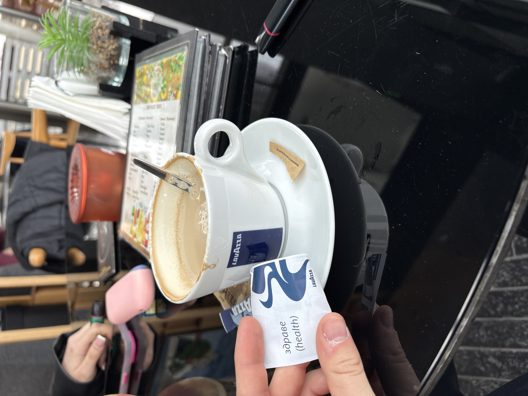 The scene depicts an almost-empty white Lavazza coffee cup tilted on its saucer, with a spoon inside, situated on a black, reflective table. A used sugar packet and an opened Lavazza-branded health sugar packet are visible. The image appears to be taken at an outdoor café or establishment, identifiable by the presence of menus, a plant in a glass container, and a table. The hand of a person holds the sugar packet. The reflections on the table, alongside the partially visible objects, add to the candid nature of the setting. The location is unknown.