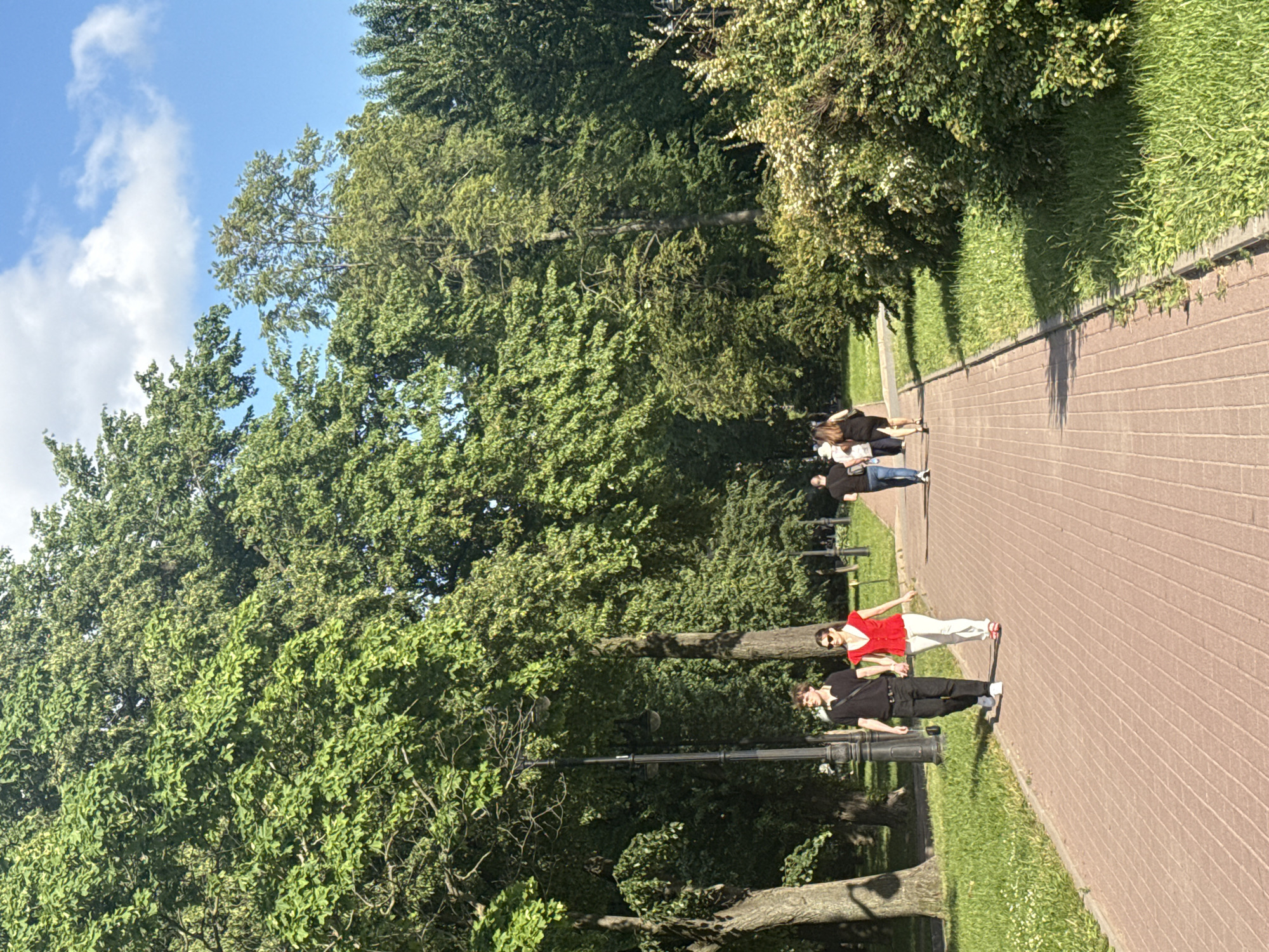 The image shows a sunny day in a park in Saint Petersburg, Russia. A paved walkway, made of red bricks, runs through the park. On the left side of the walkway, there is a grassy area with scattered white flowers and large trees. On the right side, there is a large, bushy green plant with white flowers. In the background, more trees and greenery are visible, along with a clear blue sky with a few white clouds.
Several people are walking in the park. In the foreground, a couple is walking hand-in-hand. The man is wearing a black polo shirt and black pants, and the woman is wearing a red top and white pants. Further down the walkway, a group of three people are walking away from the camera. The person in the middle is wearing a black dress, and the other two are wearing black tops and jeans or pants. Another person is walking further down the path in the distance, wearing a black top and blue jeans. A lamppost is visible on the left side of the walkway. The overall scene suggests a leisurely stroll in a well-maintained urban park.