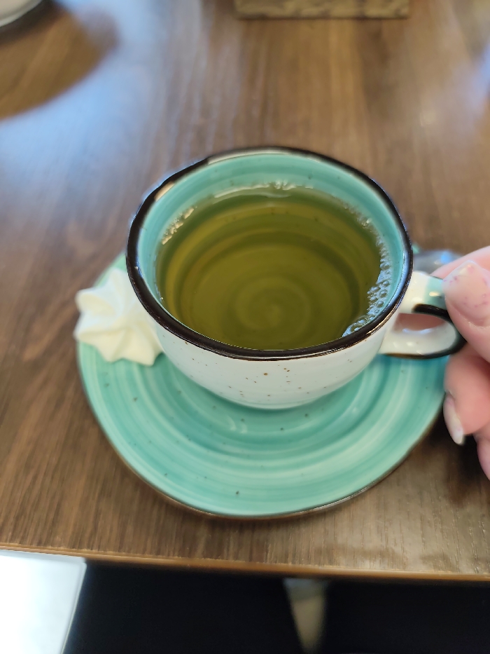 An overhead close-up shows a ceramic teacup filled with light green liquid, likely tea, on a dark brown wooden table. The liquid exhibits a swirling pattern. The cup is light beige with brown speckles, features a dark brown rim and a light turquoise interior, resting on a matching turquoise saucer with concentric rings. A small, white, star-shaped meringue is present on the saucer to the left. A human hand with manicured fingernails is seen gripping the cup's handle on the right. No specific location within Chaikovsky, Russia, is identifiable from the visual information provided.