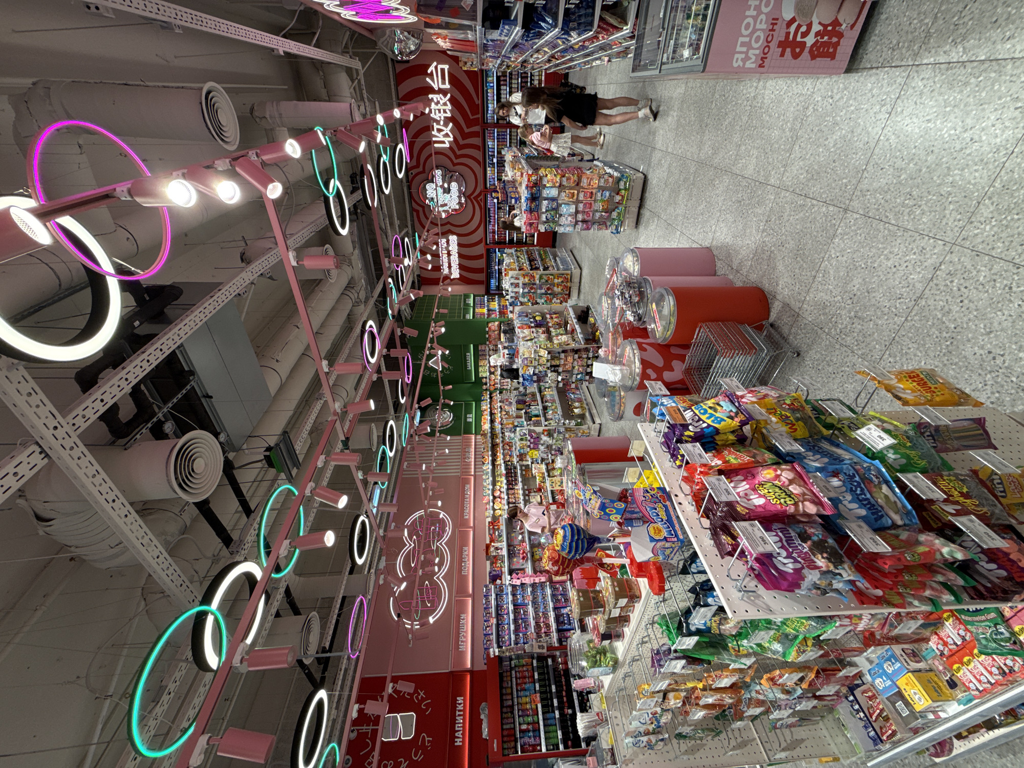 The image captures an interior view of a grocery store, presumably in Moscow, Russia, based on the Cyrillic text present. The store is brightly lit with a ceiling adorned with tubular neon lighting in varying colors of pink, white, and teal, arranged in circular and other geometric shapes. Ventilation ducts are visible on the ceiling.
Several aisles packed with various products are on display. The shelves are laden with candies, snacks, beverages, and other grocery items. Signage and promotional materials are visible in both Cyrillic and other languages, including Japanese and English. The text on some shelves includes "НАПИТКИ" (Beverages), "ИГРУШКИ" (Toys), "ПОДАРКИ" (Gifts), and "КРАСОТА И ДОМ" (Beauty and Home).
In the aisle, shoppers, including at least two children accompanied by adults, are looking at merchandise. A checkout area marked with Cyrillic characters ("收银台") is also visible in the background.