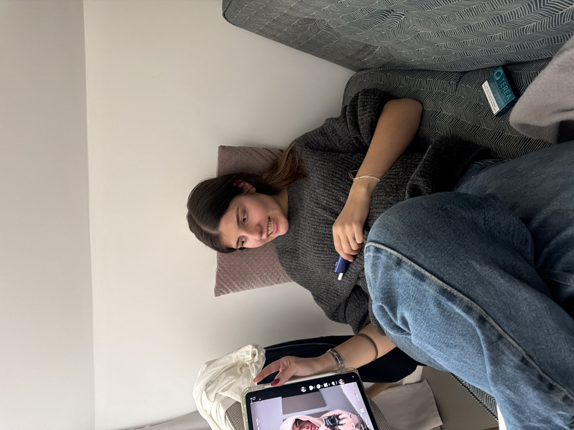 A young woman is lying on a gray couch in what appears to be an indoor setting. She is wearing a gray sweater and blue jeans. She is holding a blue pen-shaped object in one hand and has a tablet in front of her. The tablet's screen displays a picture of a person holding a camera. A box of "TEREA" is visible on the couch near her. A white wall is partially visible on the left side of the frame. The location is not discernable from the image provided. Therefore, determining if the location is in Sofia, Bulgaria, is not possible.