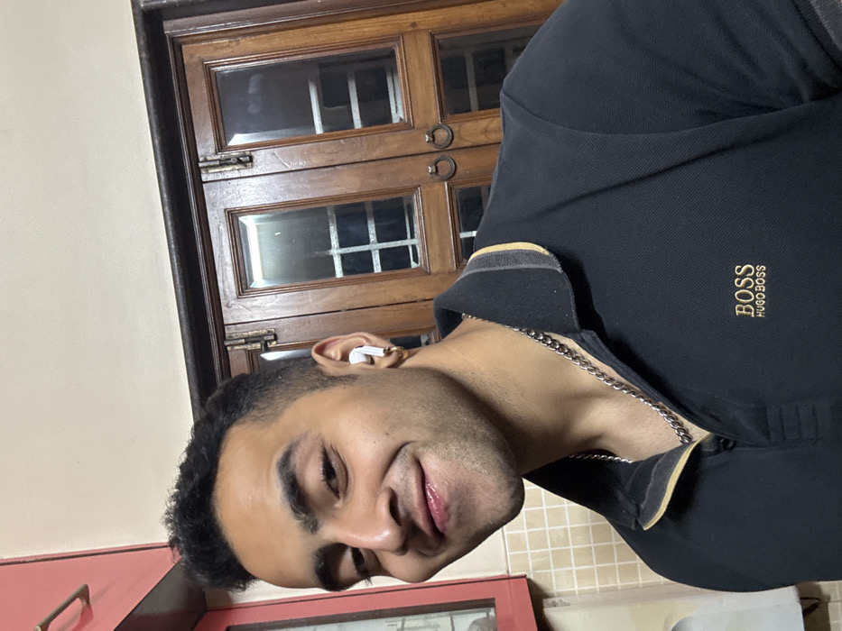 A man poses indoors wearing a black polo shirt with a gold "BOSS Hugo Boss" logo. He has an earbud in his left ear and a silver chain around his neck. A brown wooden cabinet with glass window panes and metal hardware is in the background. The photo appears to be taken at a slight angle, with a glimpse of a pink cabinet or countertop visible at the bottom left. The man smiles subtly toward the camera.