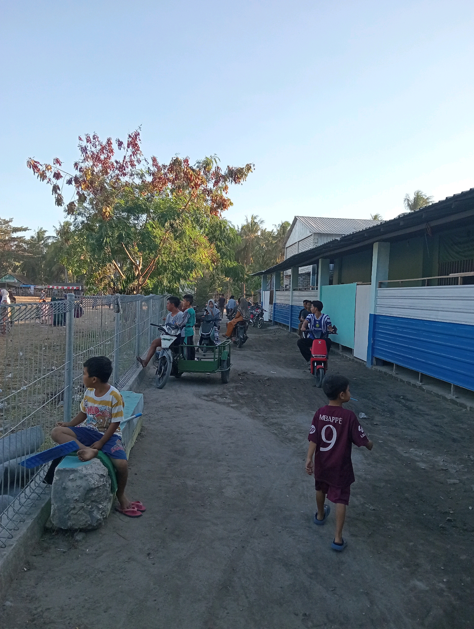 The image depicts a dusty, unpaved street in Pagerungan Kecil, Indonesia, with a group of children and adults present. On the left side of the image, a young boy wearing a striped t-shirt and blue shorts sits on a concrete ledge, looking to his left. Behind him, a metal fence encloses an area with some vegetation. Further down the street, several motorbikes are visible. One motorbike with a sidecar is ridden by a young boy, accompanied by another child, and two adults. Another young boy, wearing a maroon jersey with the number 9 and "MBAPPE" printed on the back, walks away from the camera towards a long, low building with a corrugated metal roof. This building has blue and white panels along its side. In the background, trees and other structures suggest a rural or semi-urban setting. The sky is clear and pale blue, indicating it is daytime, likely late afternoon given the shadows.