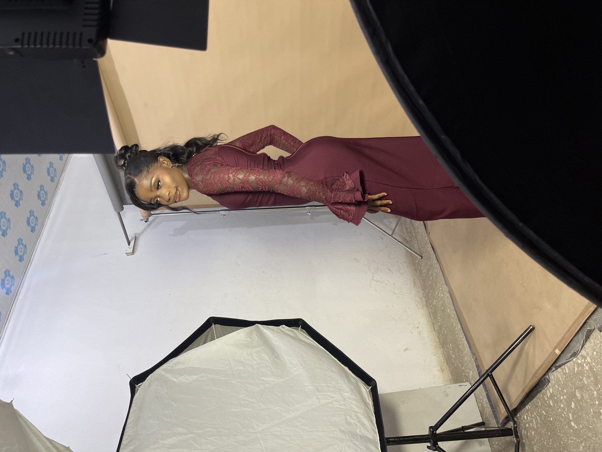 A young Black woman with her hair in a bun and styled with loose curls stands in a studio setting, posing for a photo. She is wearing a maroon, floor-length dress with lace sleeves. Behind her, a beige backdrop is visible, and to her left is a large white octagonal softbox. A camera light is positioned to her right, and the floor is a mix of concrete and a light brown mat. The ceiling has a patterned design. This scene is located in Gura Top, Nigeria.
