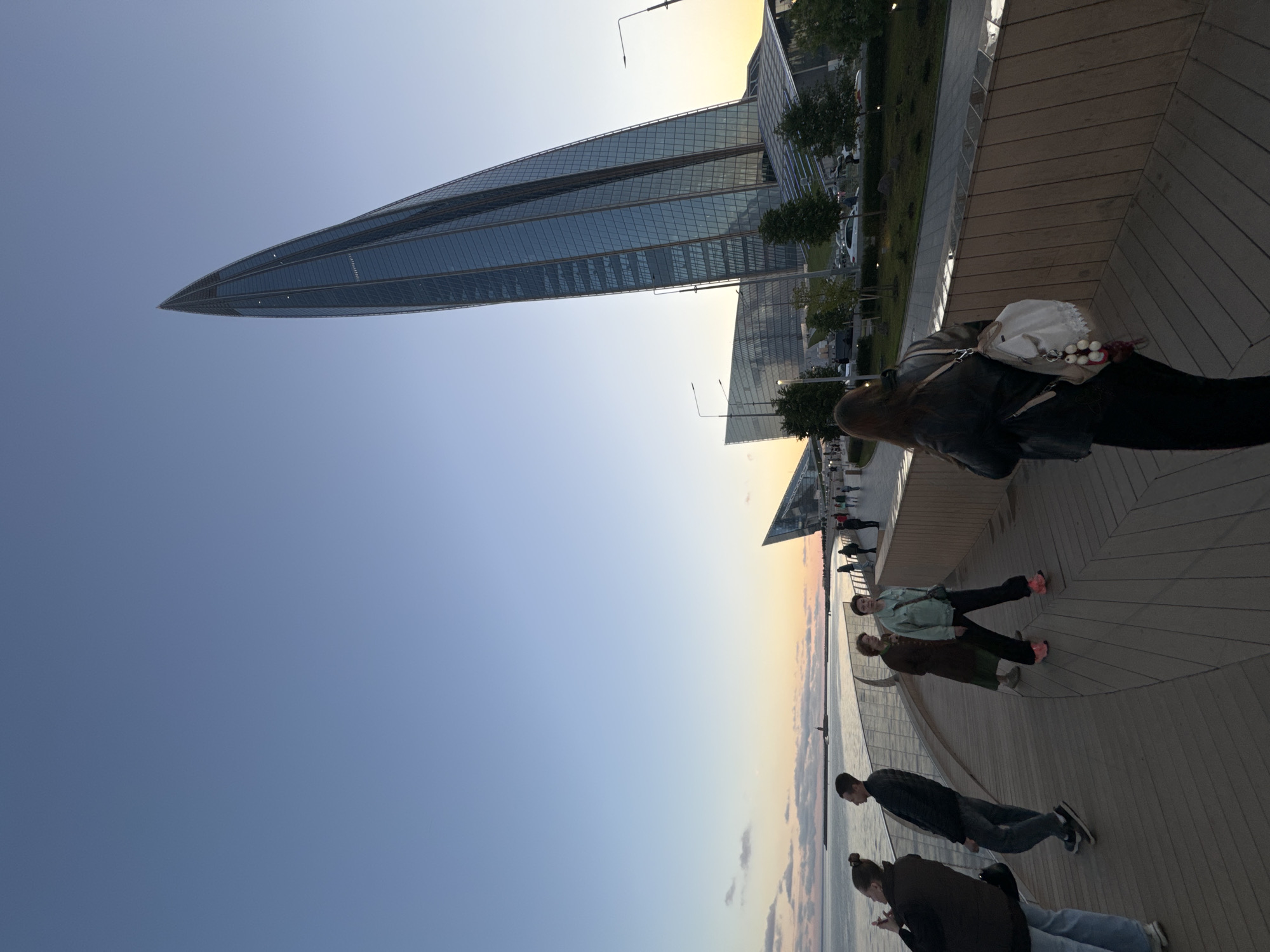 In Sinyavino, Russia, a group of people are walking on a wooden promenade beside a body of water as the sun sets. The sky is a clear gradient of blue and orange, with a few wispy clouds near the horizon. A tall, sleek skyscraper with a pointed top, made of glass and steel, dominates the background, reflecting the twilight sky. In front of it, a lower, angular building also made of glass is visible. The promenade is lined with modern railings and streetlights. Several individuals are captured mid-stride, some looking out at the water, others engaged in conversation. One person is taking a photo with their phone. The overall atmosphere is serene and urban.