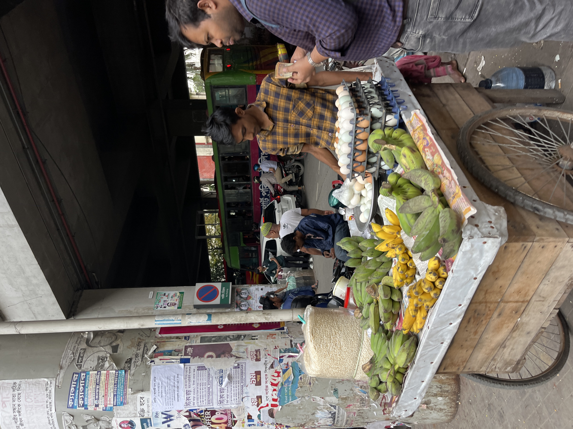 Here is a description of the image:
The image depicts a street scene in Dhaka, Bangladesh. A vendor is selling bananas and eggs from a wooden cart. The cart has a rusty wheel and is loaded with green and yellow bananas and trays of white and brown eggs. Two men are interacting with the vendor. One man in a plaid shirt appears to be paying the vendor, who is wearing a mustard-colored plaid shirt.
In the background, there is a green bus, other vehicles, and pedestrians. The scene is taking place under a flyover. A concrete pillar plastered with posters and signs is visible. The posters contain information written in Bengali. A no parking sign is also visible.