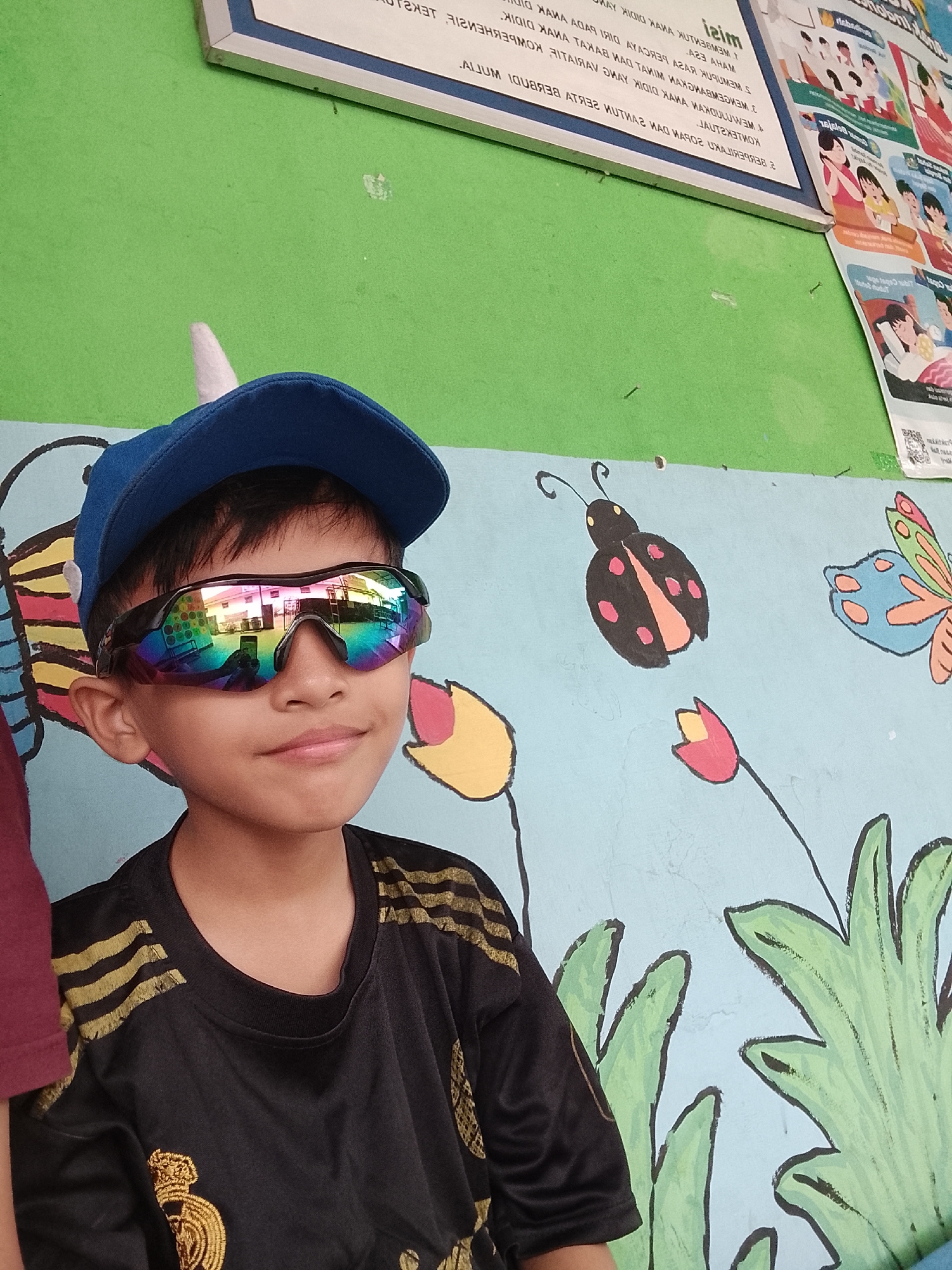 The image shows a young boy wearing a blue baseball cap with a white horn, and reflective sunglasses with a rainbow sheen, looking to his right. He is wearing a black t-shirt with gold stripes on the sleeves and a crest on the chest. He is standing in front of a light blue wall painted with a mural of a ladybug, a butterfly, a flower, and large green leaves. There is also a poster on the green wall in the background. The image appears to be taken outdoors, possibly in a school or public area in Karangkemiri, Indonesia, given the context provided by the Indonesian text on the poster. The lighting is natural, suggesting it is daytime.