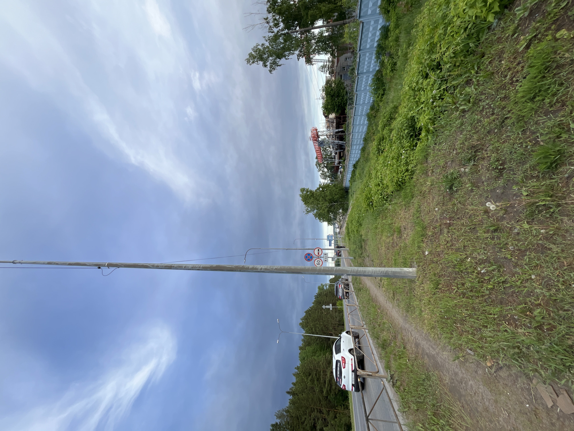 The image shows a street scene in Chaikovsky, Russia. The view is from a slightly elevated position along the side of a road. A white car is parked along a metal guardrail. Traffic moves on the road. Utility poles and traffic signs are visible, including signs indicating speed limits. On the right, grass and dense foliage cover the elevated ground beside the road, bordering a fence in the background. The sky is cloudy.