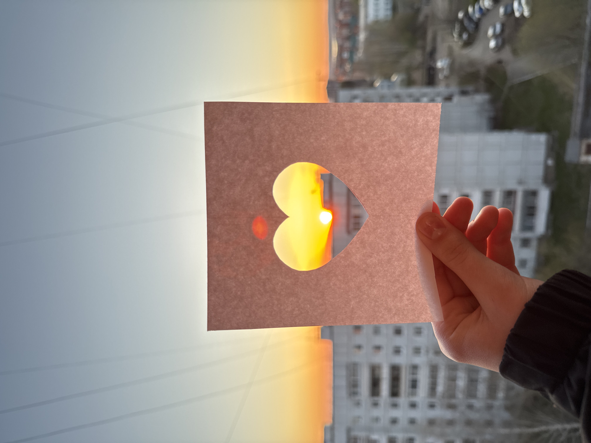 A person's hand is holding a piece of paper with a heart-shaped cutout in front of a window. The view through the window is split, with a vibrant sunset on one side and a cityscape on the other. The sunset displays a gradient of colors, from a light blue at the top to a warm orange and yellow towards the horizon. The cityscape is a mix of apartment buildings and streets, with parked cars visible. The photograph appears to be taken from inside a building, facing outwards towards the cityscape and the setting sun. The location is identified as Chaikovsky, Russia.