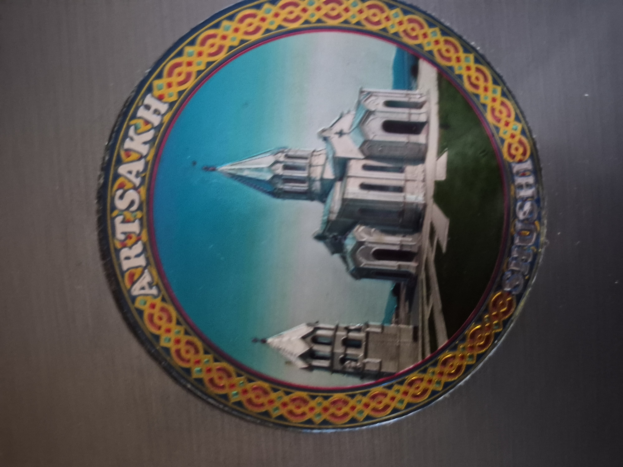 The image displays a circular magnet or badge placed on a plain, light grey surface. The magnet features a detailed depiction of a majestic, light-colored architectural complex, likely a cathedral or monastery, characterized by its prominent spires and multi-tiered structures. Two such structures are visible within the circular frame, set against a light blue sky and hints of green ground. Notably, the entire internal image of the building is rotated clockwise within the magnet, making it appear tilted.
The circular image is encircled by a decorative border adorned with an intricate, repeating pattern of red, yellow, and blue geometric motifs. Prominently displayed on this border are two pieces of text in white capital letters. The word "ARTSAKH" is visible along the top-left curve, and "SHUSHI" is seen along the bottom-right curve. The building strongly resembles the Ghazanchetsots Cathedral, a significant landmark located in Shushi, Artsakh (a region often associated with Armenia). No specific activity or event is captured, as it is a still representation on a decorative object. The scene within the magnet depicts daylight.