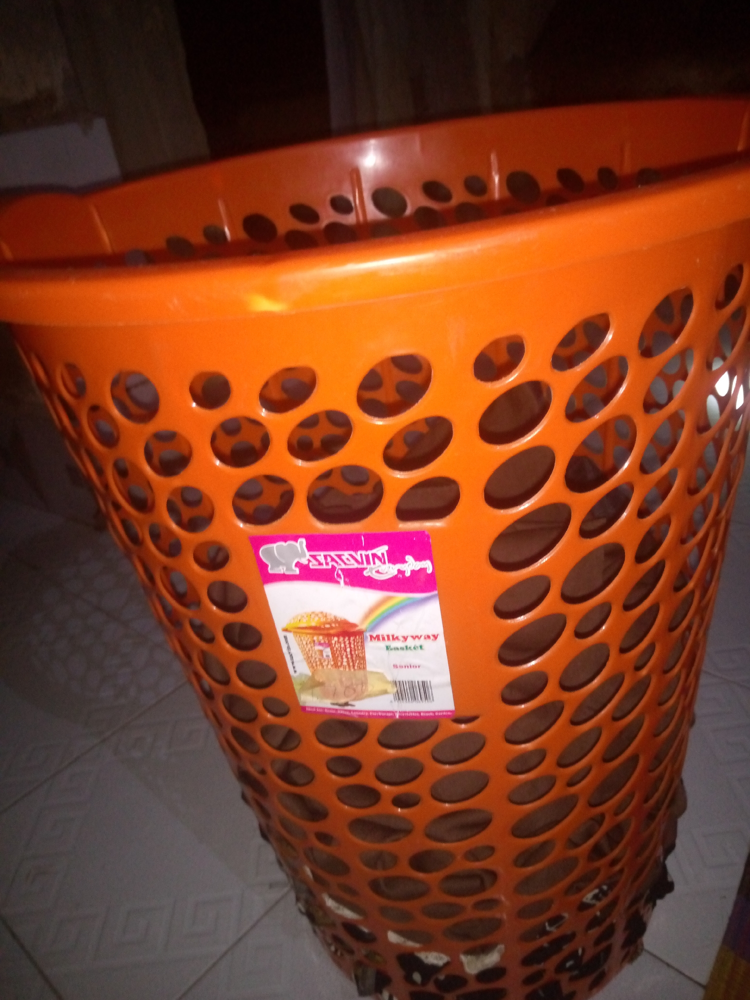 An orange plastic laundry basket is shown in the image. It is a tall, conical-shaped basket with many oval-shaped holes throughout its surface. A label is attached to the front of the basket. The label is pink and white and features an elephant graphic, along with text that reads "SAVIN" and "Milkyway Basket". The basket appears to be empty and is standing on a tiled floor. The background is dark and out of focus, with some hints of wooden structures and possibly a wall. The lighting in the image is dim.