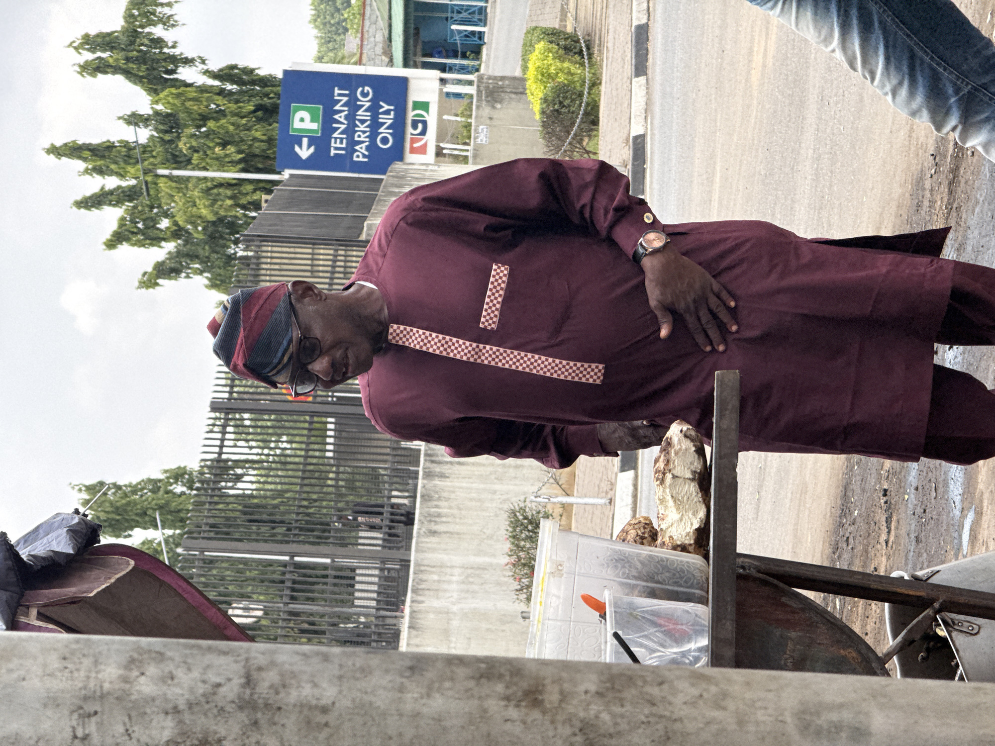 The image shows a man in a maroon tunic with checkerboard trim standing beside a food stand on a street in Abuja, Nigeria. He is wearing glasses and a patterned cap. A blue and white sign labeled "Tenant Parking Only" is visible in the background. To the right, a person wearing jeans is visible. The food stand holds containers and food items. A motorcycle is next to the food stand. The scene is set outdoors, with trees and buildings visible in the background.
