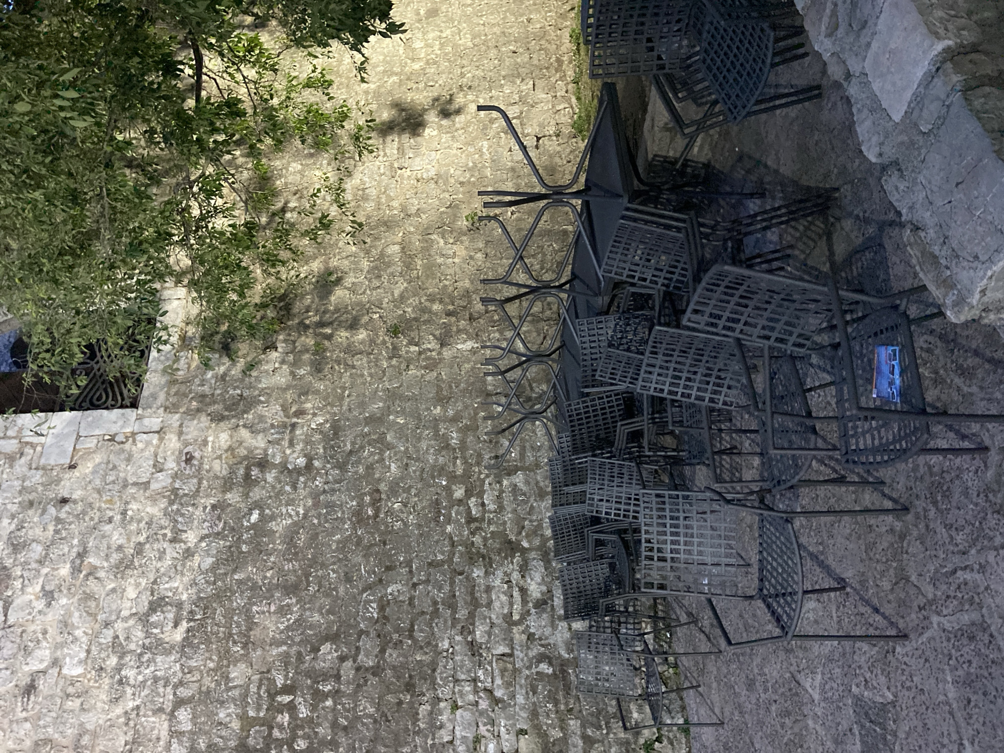 A nighttime exterior shot reveals a stack of metal chairs against a stone wall, likely in Budva, Montenegro. The chairs are stacked haphazardly, with varying angles and heights, suggesting they have been put away for the evening. The stone wall appears aged and textured, with visible mortar lines. To the left, a tree branch hangs over the scene, casting shadows on the wall. The ground surface is paved with uneven stones, adding to the old-world ambiance. No people are visible in the frame. The overall atmosphere suggests a quiet, after-hours setting.