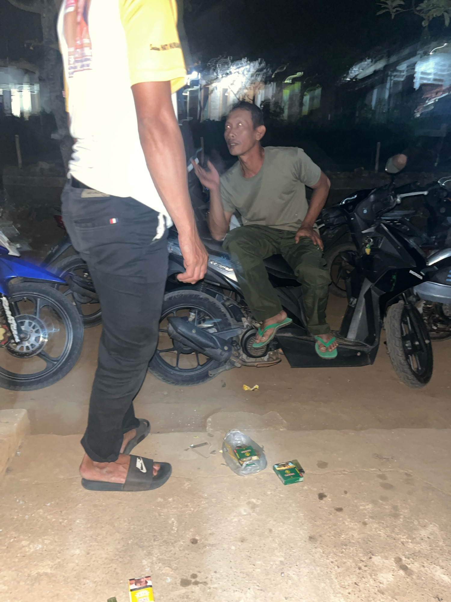 Two men are visible in the foreground and middle ground of the image. The man in the foreground is standing and facing away from the camera, wearing a yellow and white t-shirt and black pants. He is also wearing black slide sandals. The man in the middle ground is sitting on a motorcycle and facing towards the camera. He is wearing a green t-shirt and green pants, and is wearing green flip-flops. There are multiple motorcycles in the background. The scene appears to be an outdoor setting at night, possibly in the city of Kebumen, Indonesia, as indicated by the urban or semi-urban environment with buildings visible in the background and the presence of parked motorcycles, common in Indonesian street scenes. There are two packs of cigarettes on the ground in the lower right quadrant of the image. The lighting suggests artificial illumination from nearby sources.