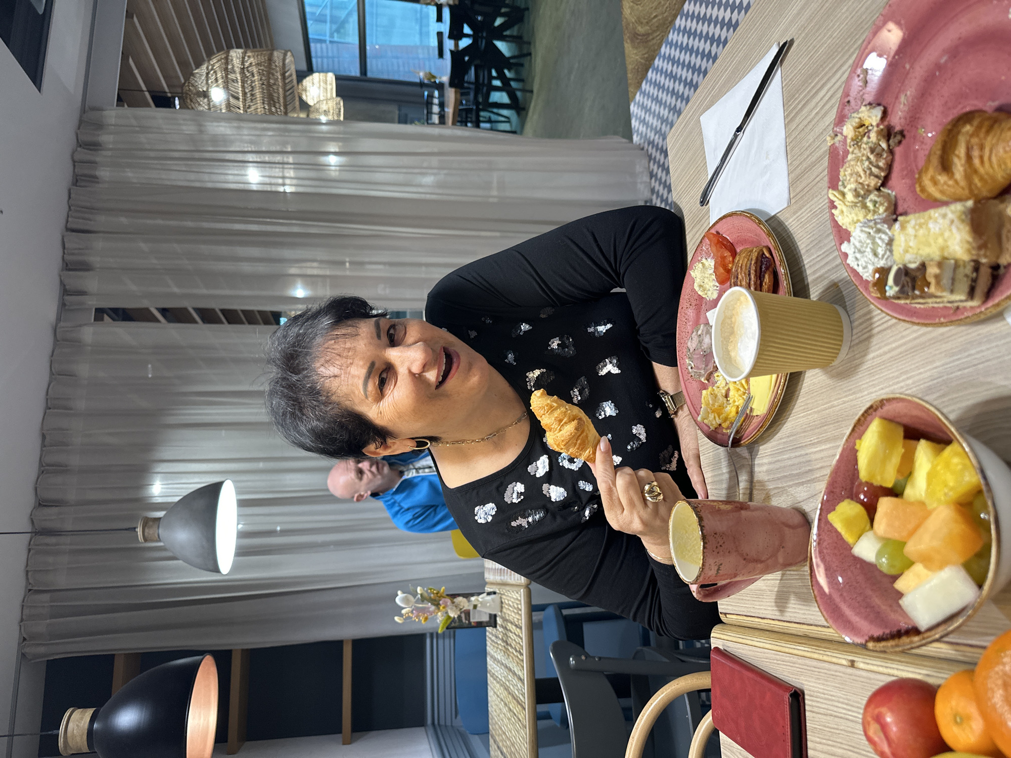 A woman is seated at a table laden with breakfast items, holding up a croissant in her right hand. She is wearing a black long-sleeved shirt with silver and white polka dots. The table contains plates with various breakfast foods, including a croissant, pastries, fruit salad in a pink bowl, and a cup of coffee. A knife and napkin are also visible on the table. Another person is seated in the background. The scene appears to be set in a restaurant in Wrocław, Poland. Woven light fixtures hang from the ceiling. The dining area is illuminated by modern hanging lights. A white curtain hangs in the background.