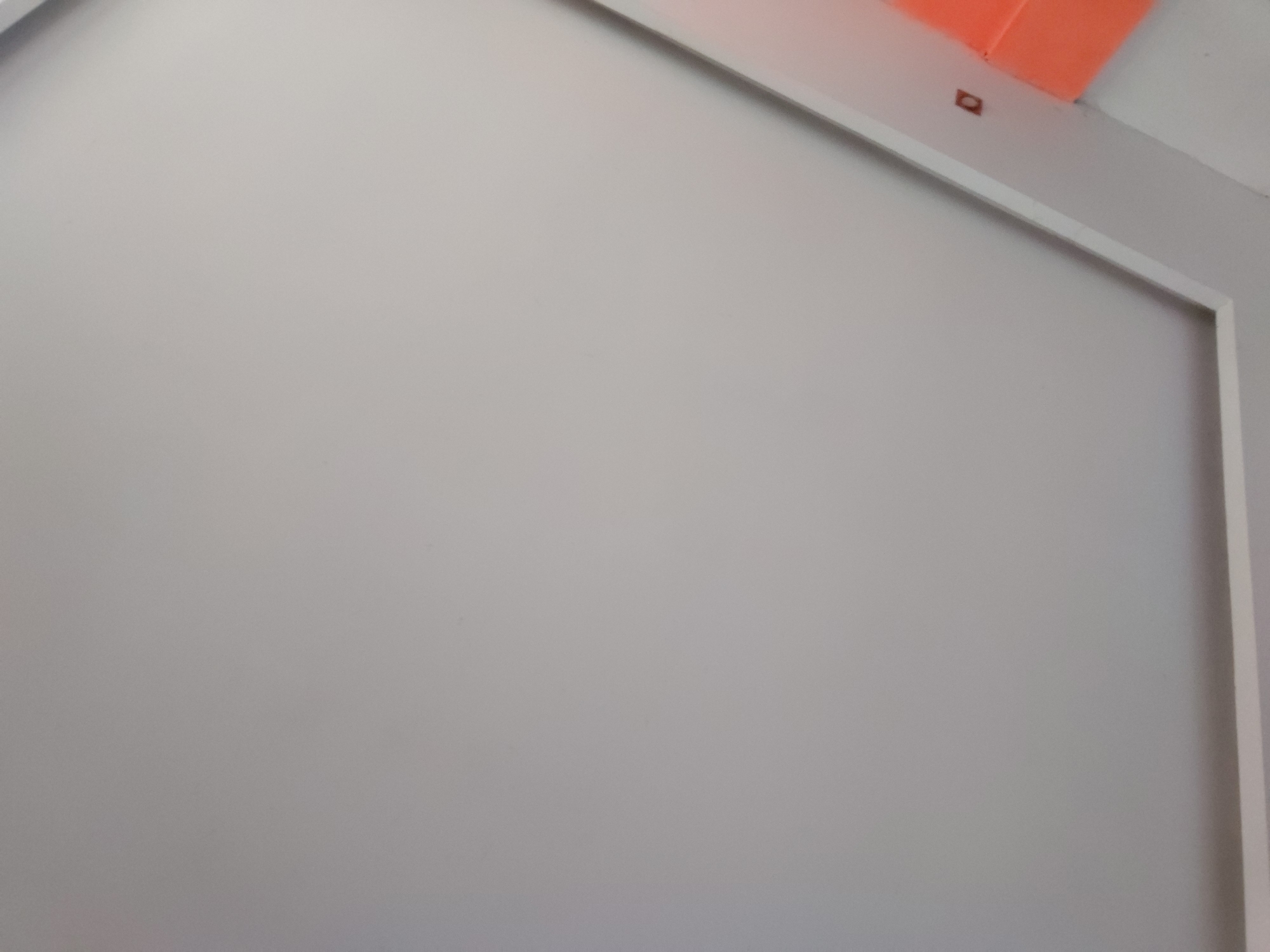 The image captures a static view of an interior space, focusing primarily on a plain, light grey wall or large surface. This grey surface dominates the foreground and extends across most of the frame. It is bordered on its right and top edges by a narrow, white architectural trim or frame, which forms an angle in the upper right section of the image.
In the top right corner, beyond the white trim, a vibrant orange object, possibly a piece of plastic or fabric, is partially visible against a lighter background. Directly below this orange object, on the white trim, there is a small, dark, rectangular or square marking or object.
No people are present, and no specific activity or event is taking place. The scene suggests a minimalist or unfinished indoor setting, likely a room or part of a structure. The lighting appears even, providing no cues about the time of day or external weather. There is no visible text within the image. The provided location context is Santa Ana, Venezuela.
