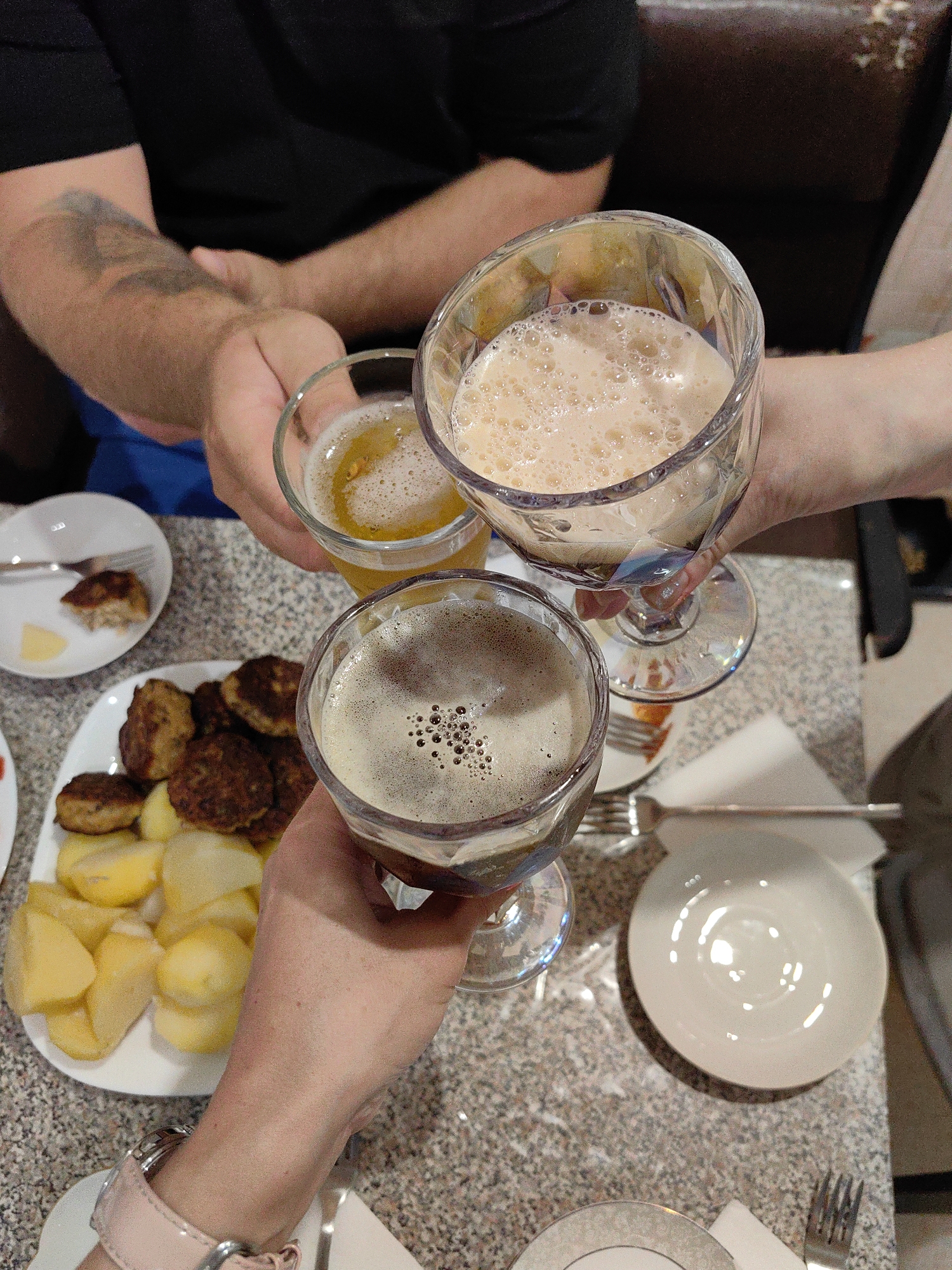 The image shows a close-up, high-angle shot of a group of people toasting with drinks in what appears to be a dining setting. The people are only partially visible, with their arms and hands holding glasses prominently featured. The hands are holding glasses filled with frothy beverages, one a lighter color with a significant amount of foam, and the other a darker color with a smaller head of foam. In the background, a plate of food is visible, containing what look like meatballs and boiled potatoes, along with a small white plate with a piece of food and a fork. The table surface has a speckled pattern. The lighting suggests an indoor environment, possibly a restaurant or home. There are no discernible landmarks or visual cues that specifically place this scene in Myadzyel, Belarus.