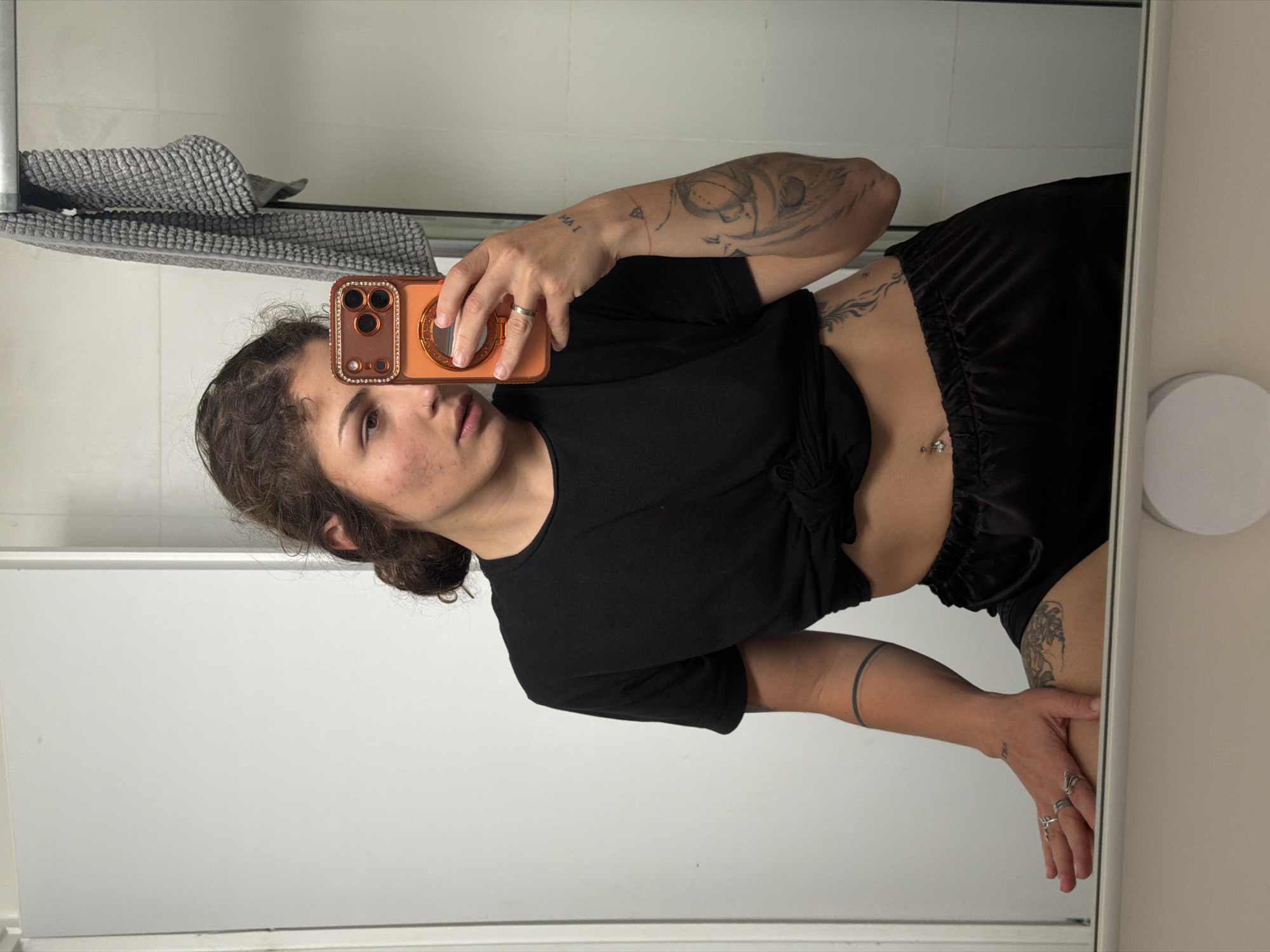 A woman takes a selfie in a bathroom mirror. She has dark hair pulled back and is wearing a black t-shirt tied at the waist and black shorts. She is holding an orange cell phone up to her face. There are tattoos on her arms and torso. The bathroom has a white wall and a towel rack with a gray towel on it. The location is unknown and there is no evidence to suggest the location is in the city of Kiryat Bialik, Israel.