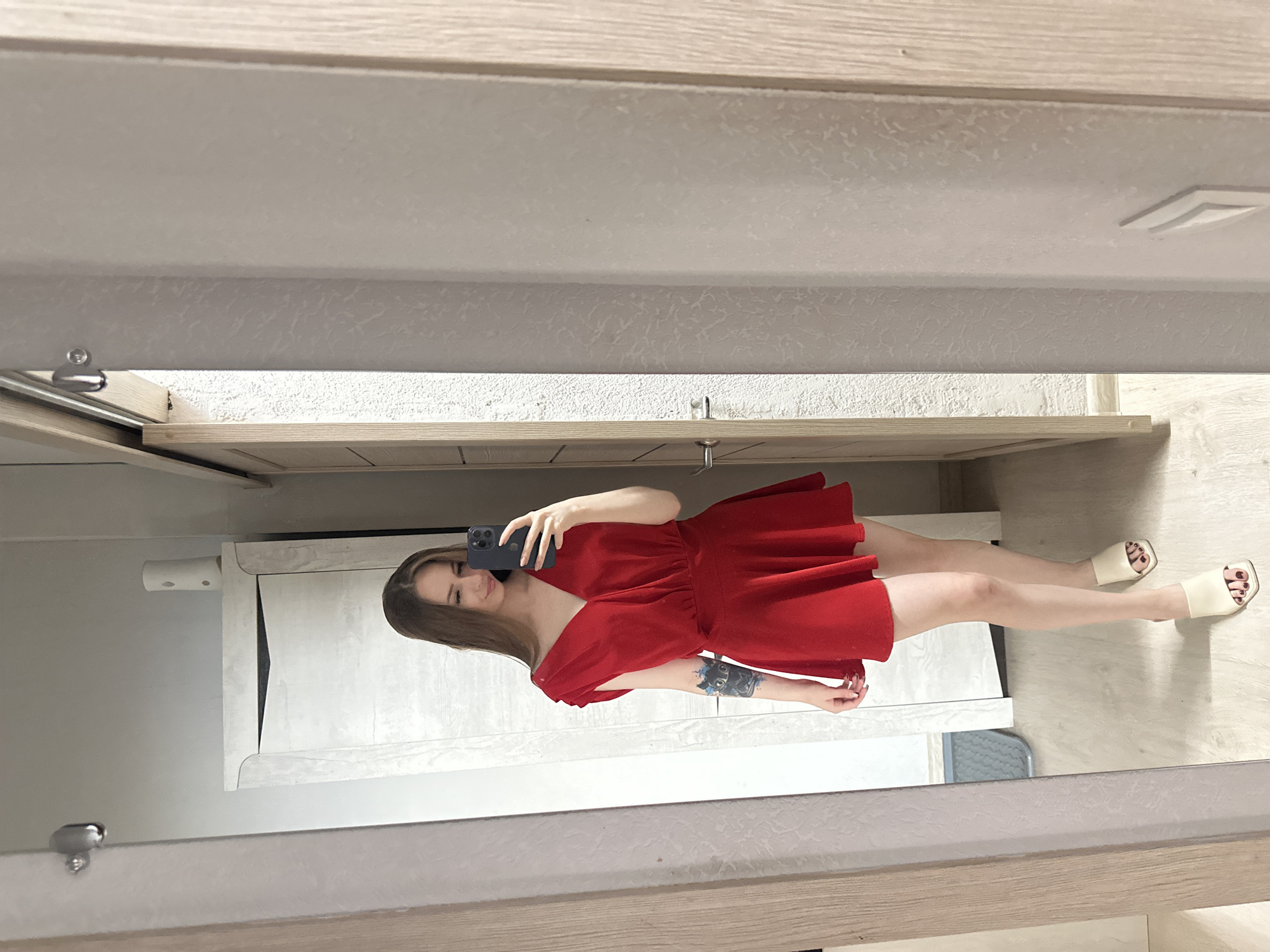 A woman is standing and taking a selfie in a mirror, wearing a red dress and white slides. The woman is facing to her right, looking at her phone and smiling. The mirror is in a room with wooden floors and doors. The woman has long brown hair and a tattoo on her left arm.