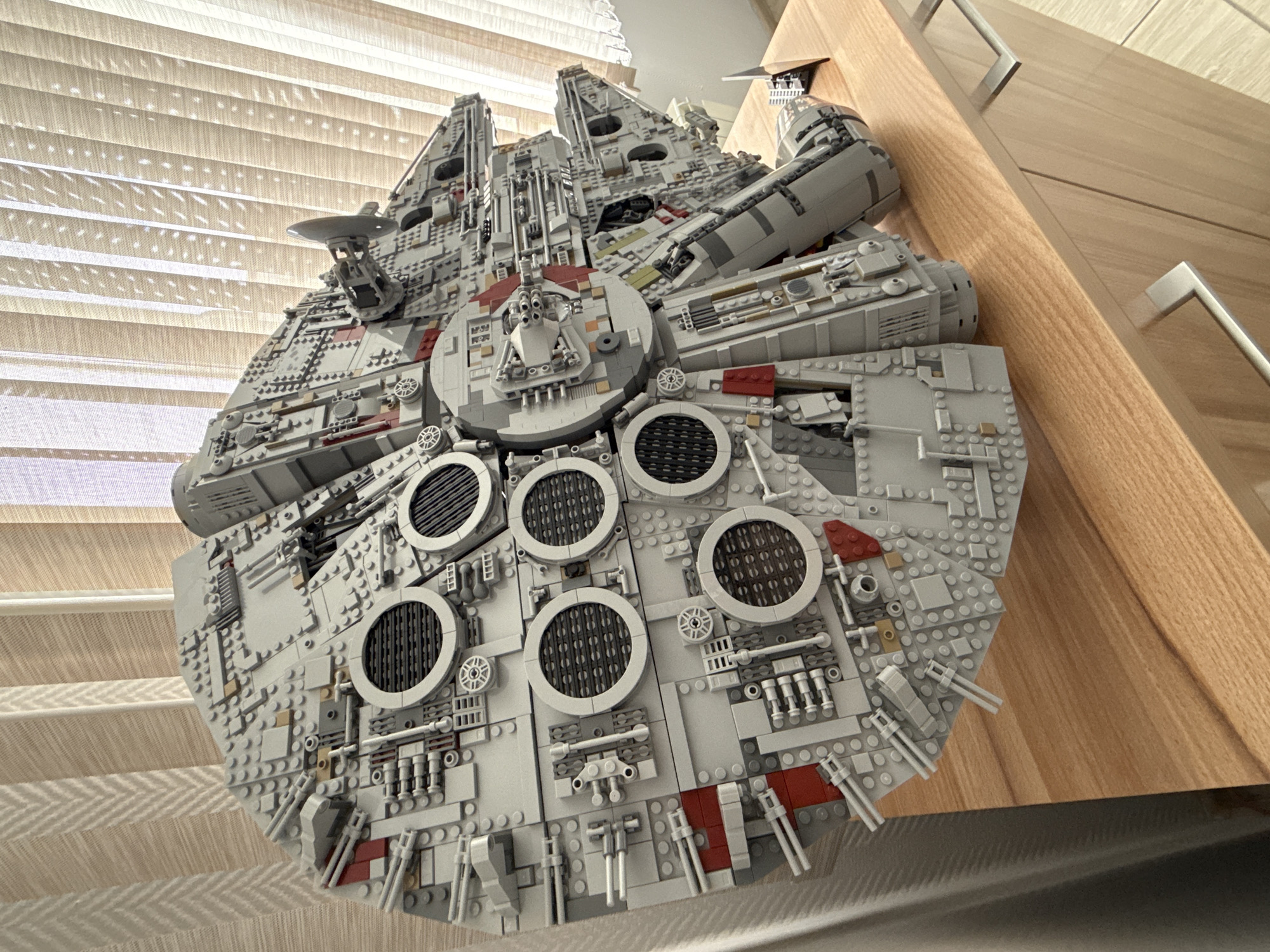 A high-angle shot depicts a detailed Lego model of the Millennium Falcon. The model rests on what appears to be a wooden surface, likely a dresser or cabinet, judging by the visible handles. The background features light-filtering blinds. The image provides no indication that the location is Krasnoyarsk, Russia, nor does it contain any information about people or actions.