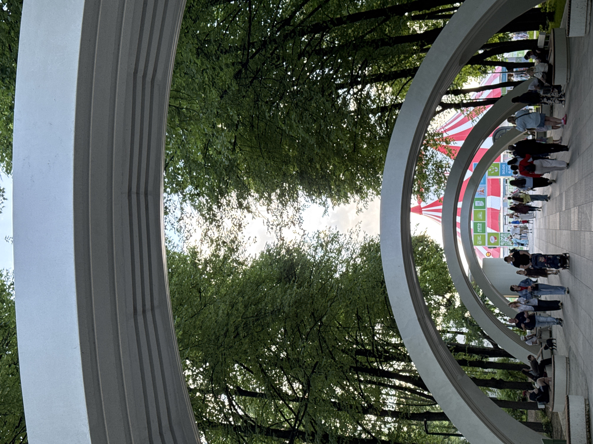 The image depicts a series of arched concrete structures in a park-like setting. Through the arches, trees are visible, and in the distance, a red-and-white striped tent can be seen. On the right side of the image, people are walking along a paved path. The image appears to be taken in Moscow, Russia.