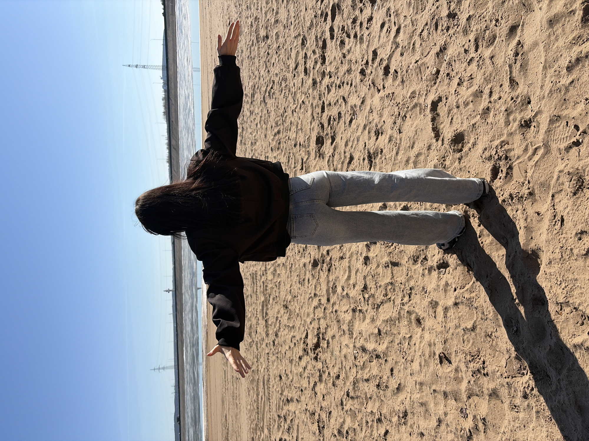 A full-length, vertically oriented photograph captures a person standing on a sandy shore with their back to the viewer, arms outstretched horizontally. The individual has long, dark hair and is wearing a dark hooded top or jacket and light-wash blue jeans. They are looking out towards a large body of water under a clear, bright blue sky.
The scene depicts a sunny day, likely late morning or afternoon, as evidenced by the distinct, long shadows cast on the sand. The sandy ground is textured with footprints and small undulations. To the left of the person, the calm surface of the water reflects the bright sky, showing gentle ripples.
In the distance, across the water, the horizon reveals several tall electricity pylons and power lines. The person's pose, with arms outstretched, evokes a sense of freedom or expansiveness, contemplating the vastness of the water and sky. The weather is clear and sunny. No visible text is present in the image. The setting is a sandy coastline in Noviy, Russia.