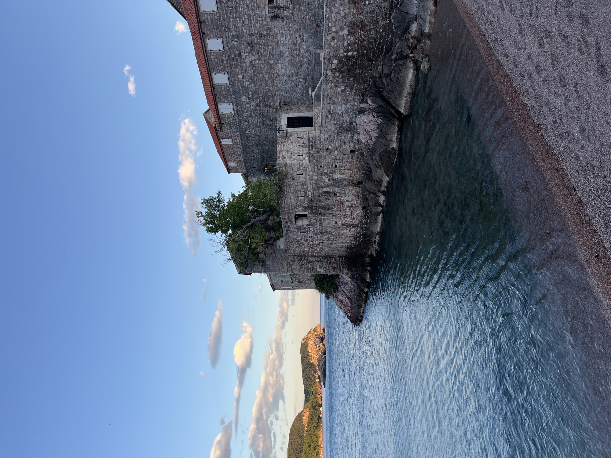 This image captures a picturesque coastal scene, strongly resembling Sveti Stefan, Montenegro. A prominent, multi-story stone building with red tiled roofs and numerous windows, many fitted with white shutters, stands tall on a rocky outcrop. The architecture suggests historical significance, with rough-hewn stone walls and an arched entryway featuring dark doors and two outdoor lanterns on a lower level. A small tree or shrub is seen growing from the side of the building, nestled into the rocky structure.
To the right of the building, a calm expanse of deep blue-green water stretches out, meeting a narrow, dark pebble beach or shoreline. Gentle ripples are visible on the water's surface, reflecting the clear sky. In the background, across the water, a series of undulating hills and mountains rise, covered in rich greenery and bathed in the warm, golden light of what appears to be late afternoon or early evening. The sky overhead is a vibrant blue, adorned with scattered, fluffy white clouds. The overall atmosphere is one of serenity and timeless beauty, blending ancient architecture with a tranquil natural landscape. There is no visible human activity or text within the frame.