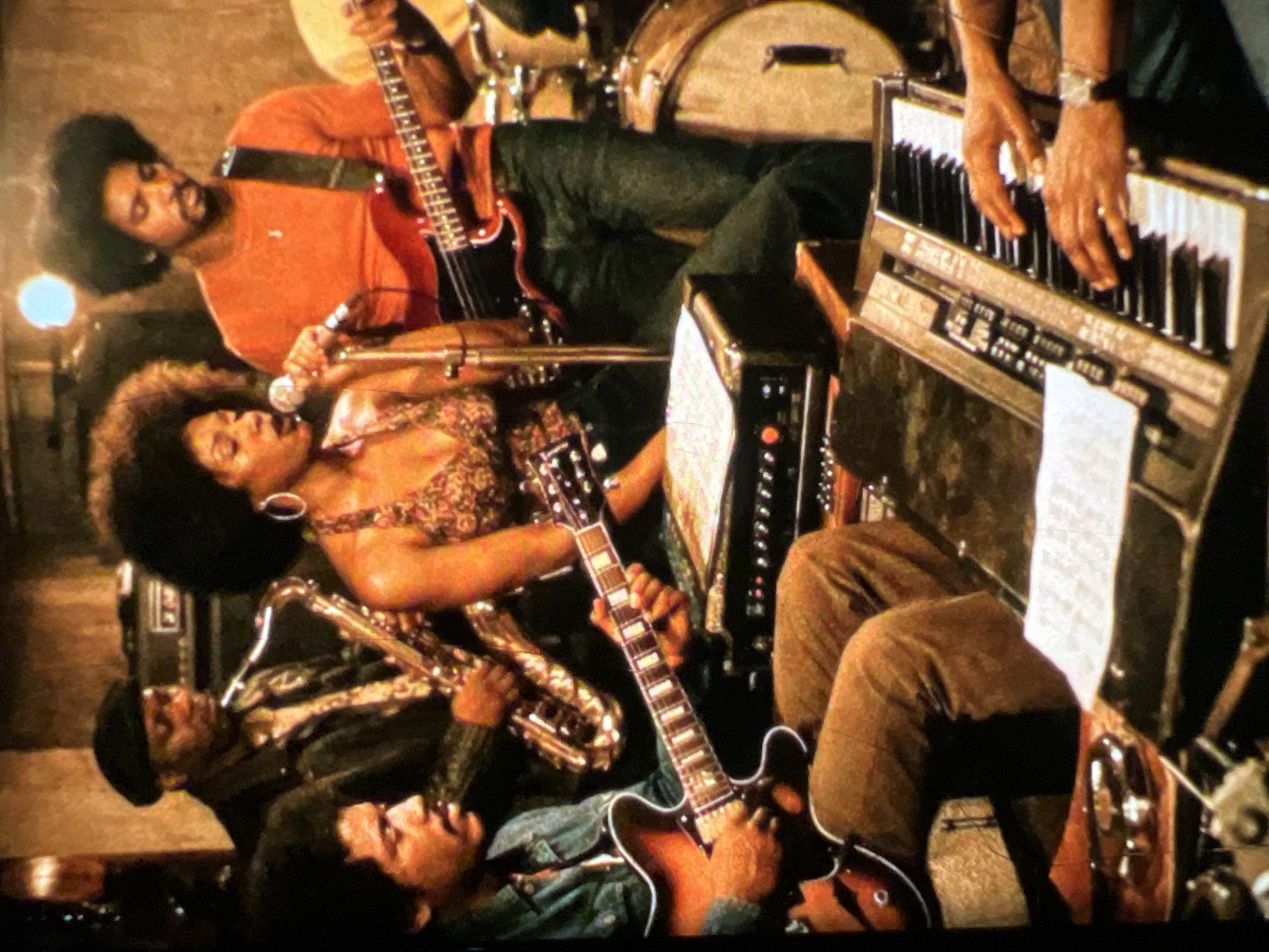 The image depicts a band performing in a dimly lit indoor space. Five individuals are visible, actively engaged in playing musical instruments. In the foreground, a person is playing a keyboard, with their hands positioned over the keys. To the left, another person is playing a guitar. In the background, a vocalist is singing into a microphone, accompanied by a saxophonist and a bassist who is also playing a guitar. The environment appears to be a music venue or studio, with musical equipment such as amplifiers and drums also present. The overall atmosphere suggests a live music performance. The image does not contain any specific visual cues that would allow for the determination of the location within the city of Lagos, Nigeria.