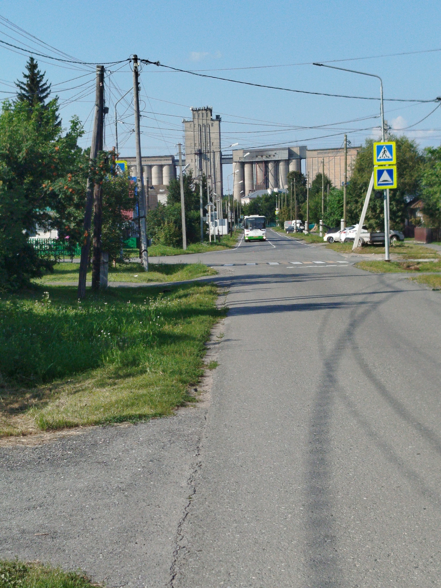 The image depicts a street scene in the city of Zavodoukovsk, Russia, featuring a road with a pedestrian crossing. In the background, there are large industrial buildings, likely grain elevators or silos, with a clear blue sky overhead. A green and white bus is visible on the road, approaching the pedestrian crossing. Two yellow and blue traffic signs are mounted on a pole on the right side of the image, one indicating a pedestrian crossing and the other a warning sign with a triangle. Power lines are strung across the top of the frame, supported by wooden poles. The sides of the road are lined with grass and some trees. Several cars are parked or driving in the distance. The overall impression is of a functional, somewhat industrial, urban environment on a clear day.
