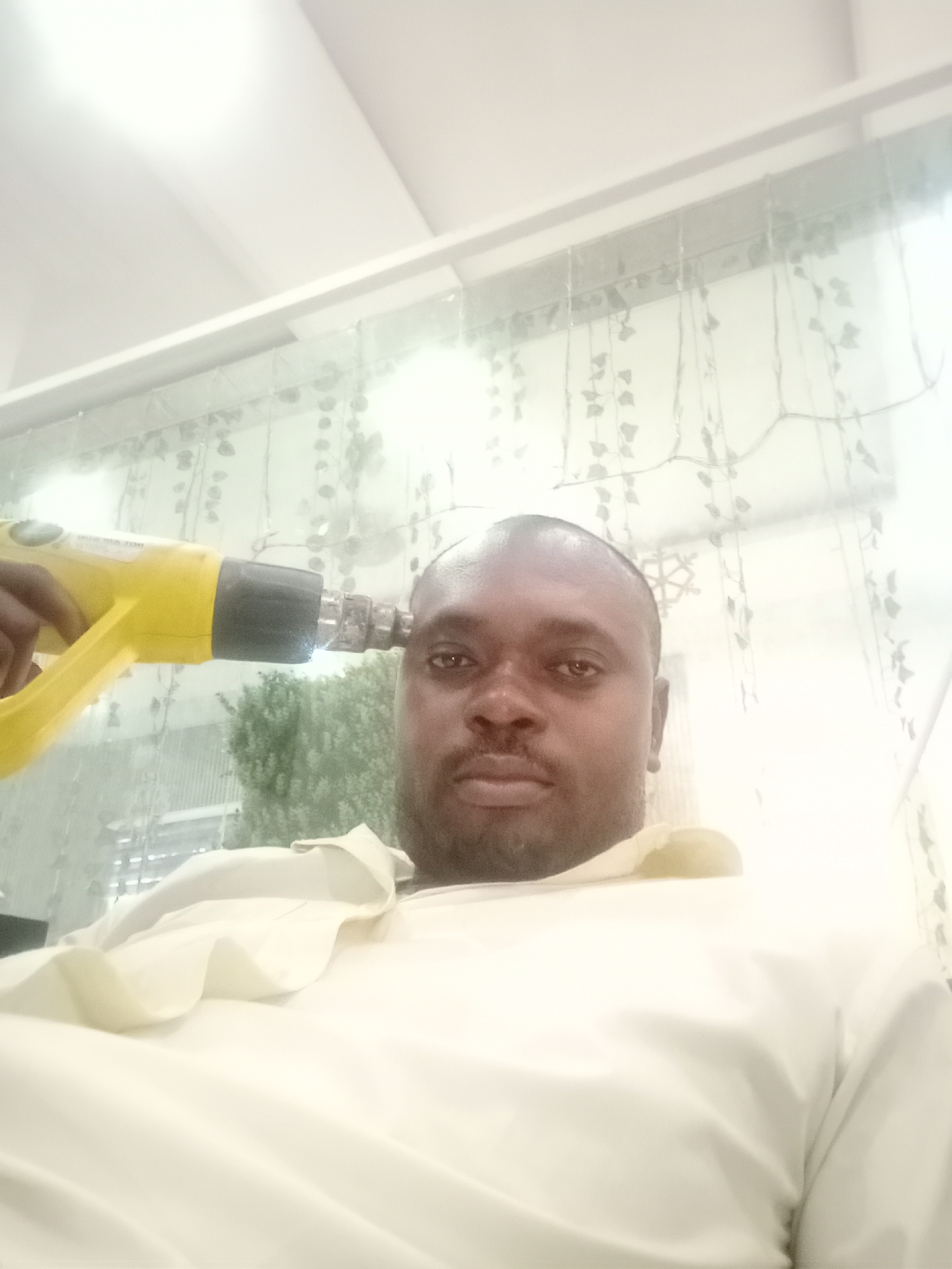 The image depicts a dark-skinned man with a beard, facing forward, with a yellow heat gun pointed at his head. He is wearing a light-colored garment. The background features a curtain or partition with hanging decorative elements resembling leaves, and hints of greenery visible through it. The setting appears to be indoors. There is no way to verify the location in Gwarinpa, Nigeria.
