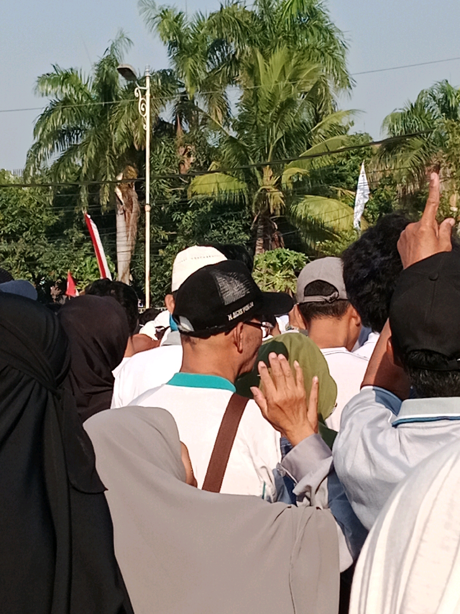 The photograph captures a crowd of people gathered outdoors, with lush palm trees and greenery forming the backdrop. Several individuals are visible in the foreground, most of whom are facing away from the camera or in profile. They are dressed in casual attire, with some wearing head coverings. One person in a black baseball cap is looking towards the left, while another in a white cap is slightly behind and to their right. A person in a light grey hijab is positioned in the lower part of the frame, partially obscuring another individual. The environment appears to be a public space, possibly a park or a street, under a bright sky. The overall scene suggests a community gathering or event. The image was taken in the city of Surakarta, Indonesia.