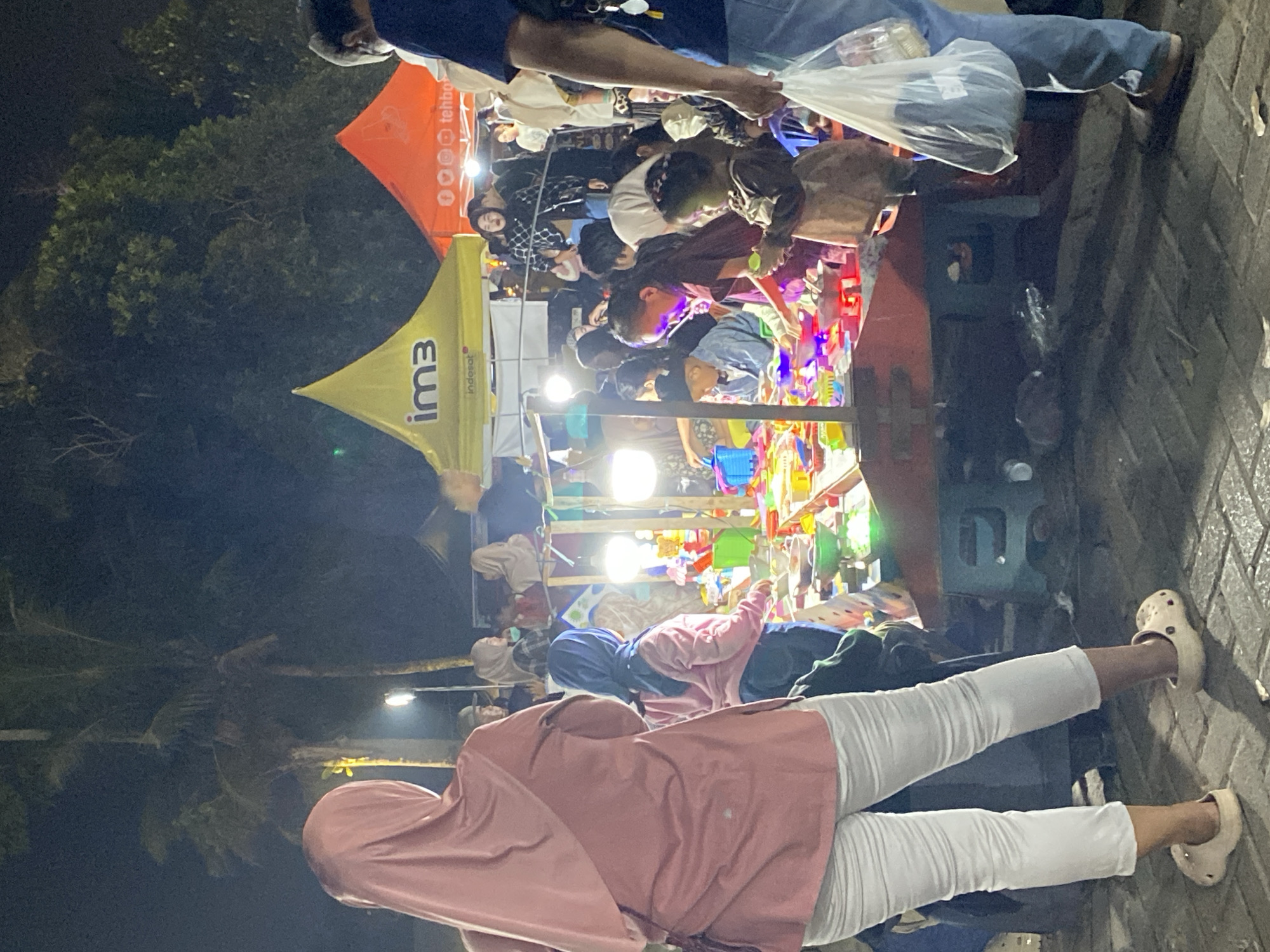 This image depicts a bustling night market scene in Banjarmasin, Indonesia, likely on a paved street with a cobbled texture. The foreground is dominated by a woman from the back, wearing a light pink hijab and white capri pants, with light beige Croc-style shoes. She is standing on the left, facing a colorful stall filled with various items, possibly toys or trinkets, illuminated by bright lights.
The stall is the central focus, a long, red table laden with goods. A variety of people, including adults and children, are gathered around it, browsing and interacting. Among them, a child in a brown dress is holding a small yellow object. Another child in a blue hijab and pink top is looking at the items on the stall. Several other individuals are visible in the background, their faces partially obscured by the crowd and the dim lighting.
Behind the stall, two large tents are set up, one bright yellow with the logo "im3" and "indosat", and another orange tent with circular graphics and text that appears to read "tehbotol". Palm trees and other greenery are visible in the darker background, suggesting an outdoor setting, possibly near a park or a wide street. The overall atmosphere is lively and crowded, indicative of a popular local gathering. The lighting is a mix of artificial sources from the stall and tents, creating a vibrant, nighttime ambiance.