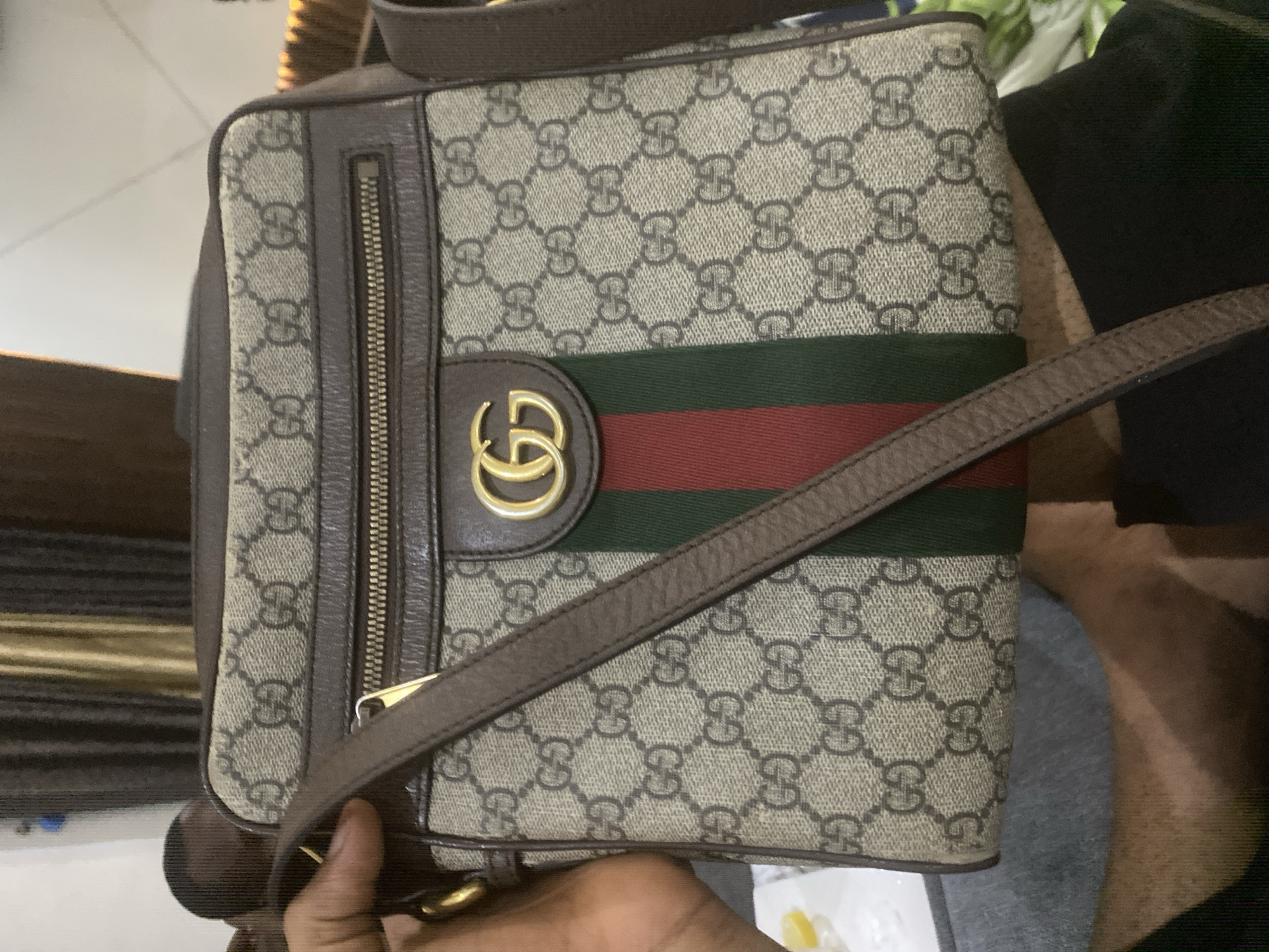 A brown and beige Gucci handbag is the central subject of the image, held by a person whose hand is visible at the bottom. The handbag features the iconic Gucci GG Supreme canvas pattern in beige with dark brown interlocking "GG" monograms. It is trimmed with dark brown leather along its edges. A prominent green and red Web stripe runs horizontally across the front of the bag, with a gold-toned interlocking "GG" logo (Marmont style) positioned centrally on the stripe.
To the left of the central panel, a vertical zippered pocket is visible, featuring a gold-toned zipper and pull. A thin, dark brown leather shoulder strap extends from the bag, partially held by the person's hand. The texture of the canvas, the leather grain, and the metallic sheen of the hardware are discernible.
The activity taking place appears to be the presentation or display of the handbag, possibly for a photograph. The setting is indoors, with a blurred background that includes light-colored surfaces and some darker, indistinct objects, along with a grey fabric in the lower right, possibly clothing. The lighting is bright, suggesting daytime. The "GG" monogram is the most prominent visible text.