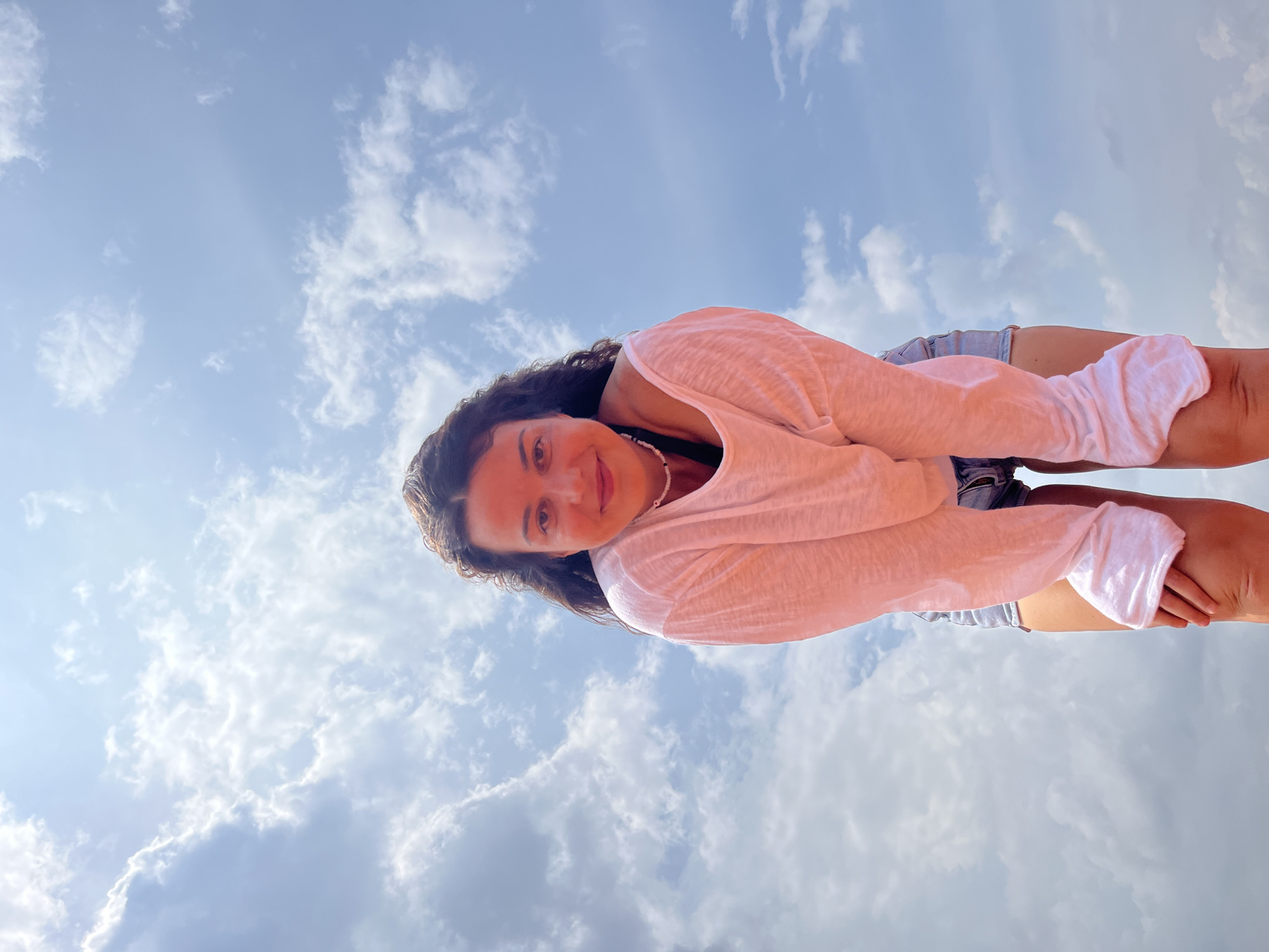 A medium shot captures a woman from a low angle, looking directly at the camera with a slight smile. She has long, dark, curly hair and is wearing a loose-fitting, white, long-sleeved shirt with the sleeves pulled down and a pair of denim shorts. A thin, beaded necklace is visible around her neck. Her hands are resting on her knees, and she is bent forward slightly. The background is a bright blue sky filled with white, puffy clouds, suggesting an outdoor setting, possibly in Mui Ne, Vietnam, given the appearance of the sky and the woman's attire, which is suitable for a tropical climate. The lighting indicates it is daytime, with sunlight filtering through the clouds.