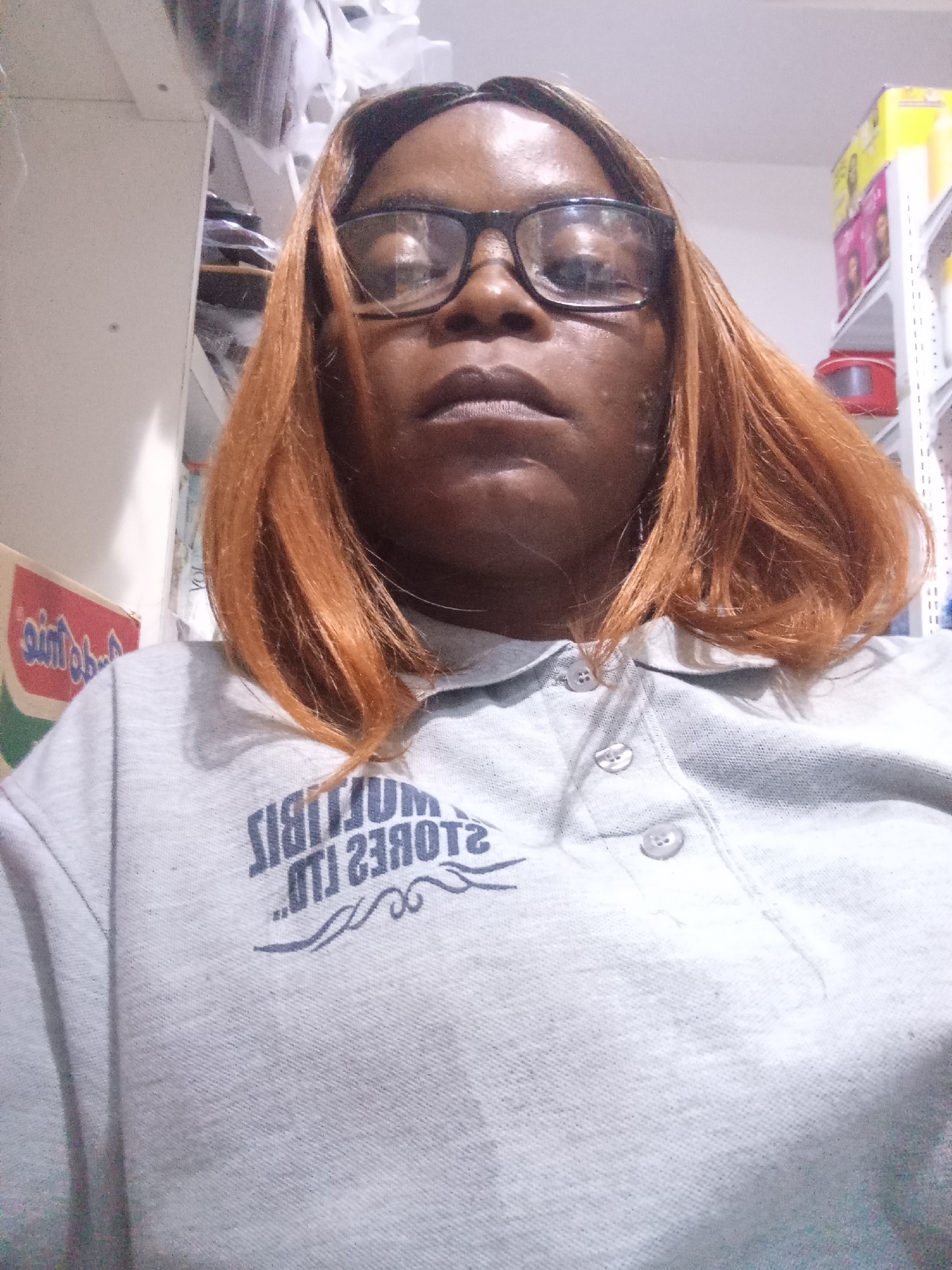 A medium shot shows a Black person with ginger-colored hair, wearing glasses and a light grey polo shirt with a dark blue logo that appears to say "MULTIBIL STORES LTD" in reverse. The person is looking directly at the camera with a neutral expression. The background is blurred but suggests an indoor environment, possibly a retail setting with shelves stocked with items. The lighting is artificial and appears to be from overhead. The image appears to be a selfie taken in Howayi, Nigeria.