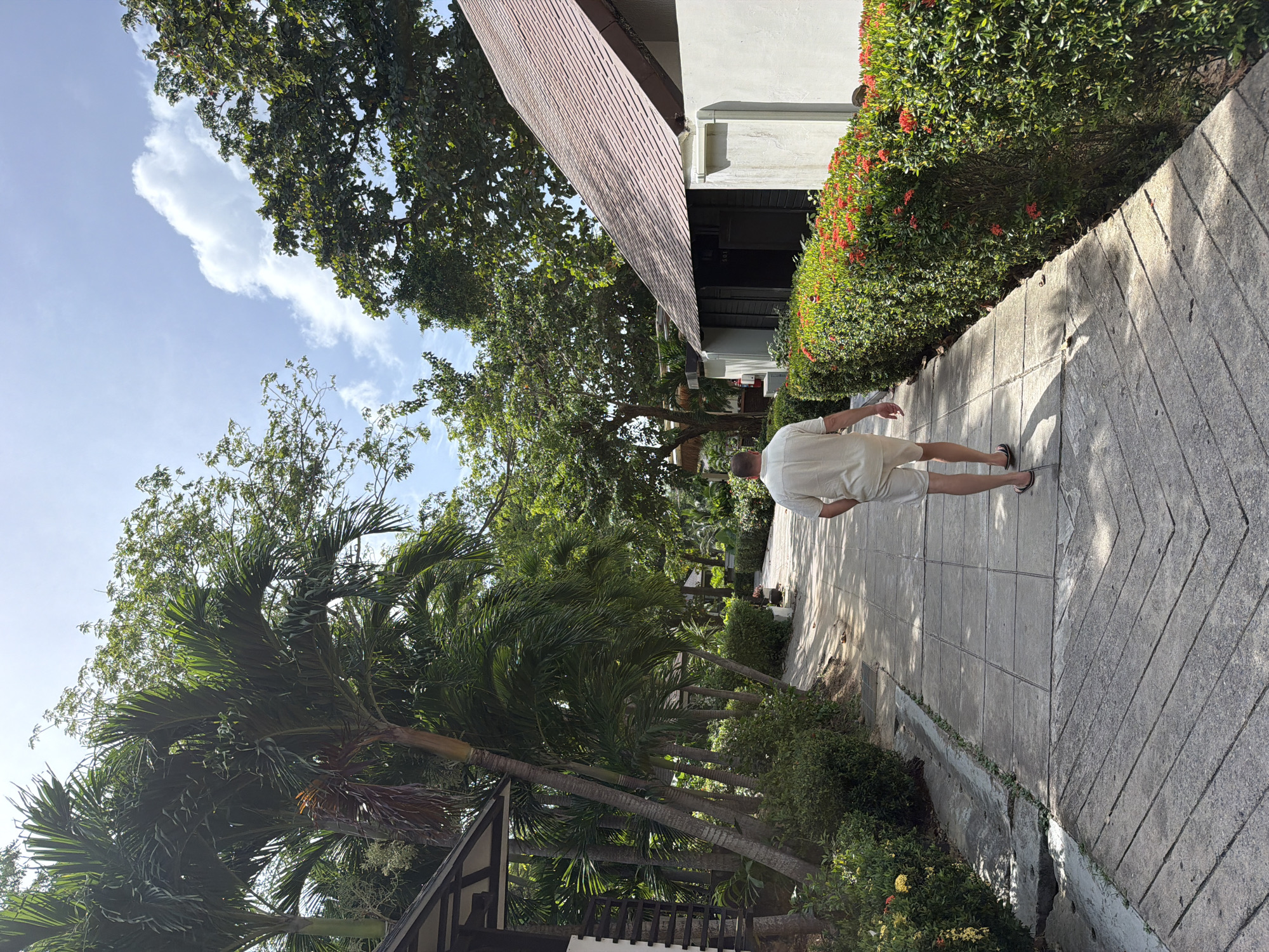A man walks away from the camera on a paved pathway bordered by lush green bushes with red flowers and palm trees. Buildings with pitched roofs are visible on either side of the pathway. The sky is blue with scattered white clouds. This appears to be a resort or hotel in Ao Nang, Thailand.