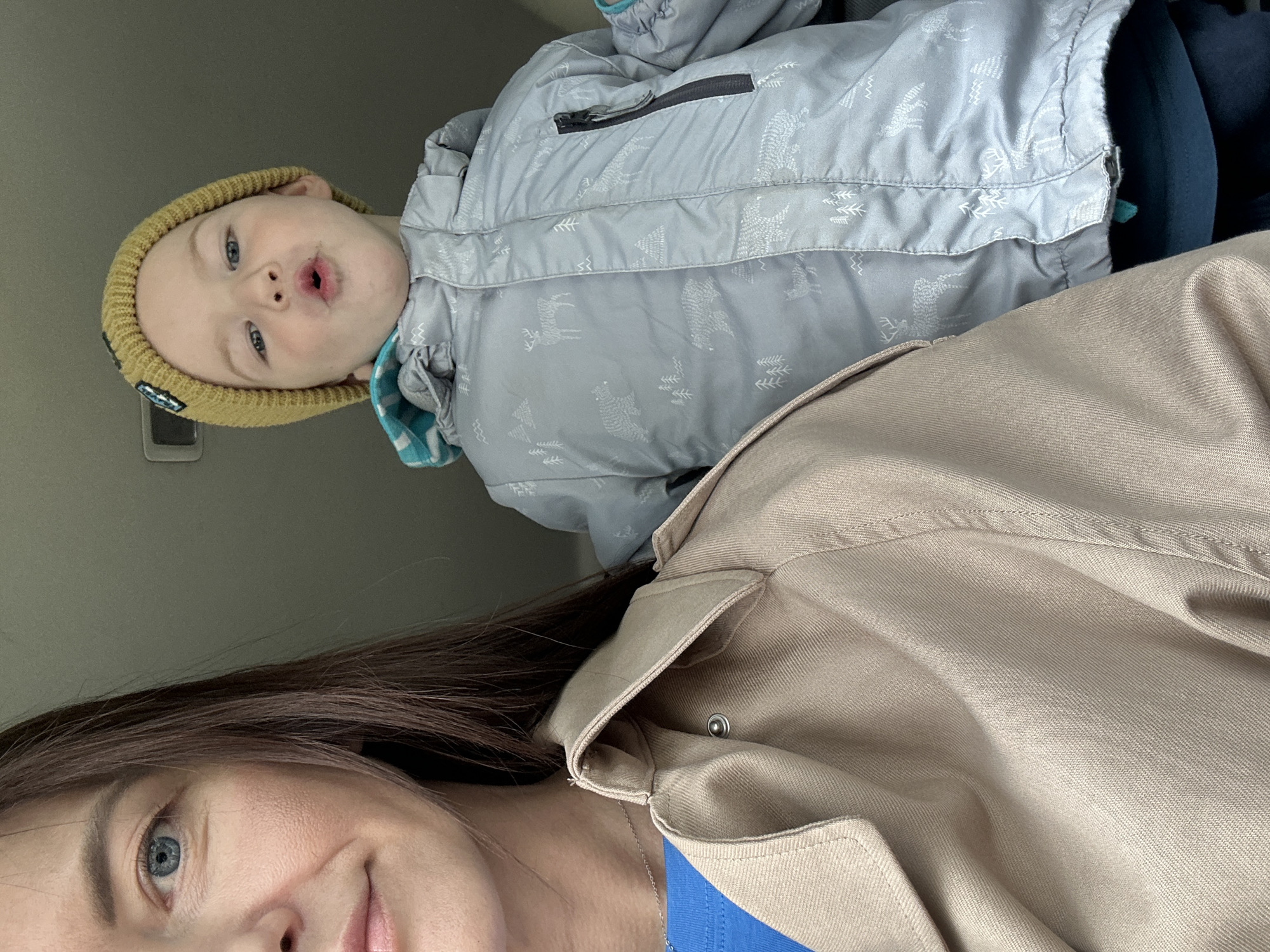 A woman and a young child are visible in a close-up, tilted selfie-style image. The woman has fair skin, brown hair, and blue eyes, and is wearing a beige jacket over a blue top. The child, who appears to be a toddler, is wearing a light gray winter coat with a pattern of trees and reindeer, along with a yellow knit cap. The background appears to be the interior of a car, with a gray interior panel visible. The location in the city of Chaikovsky, Russia is not discernible from the image.