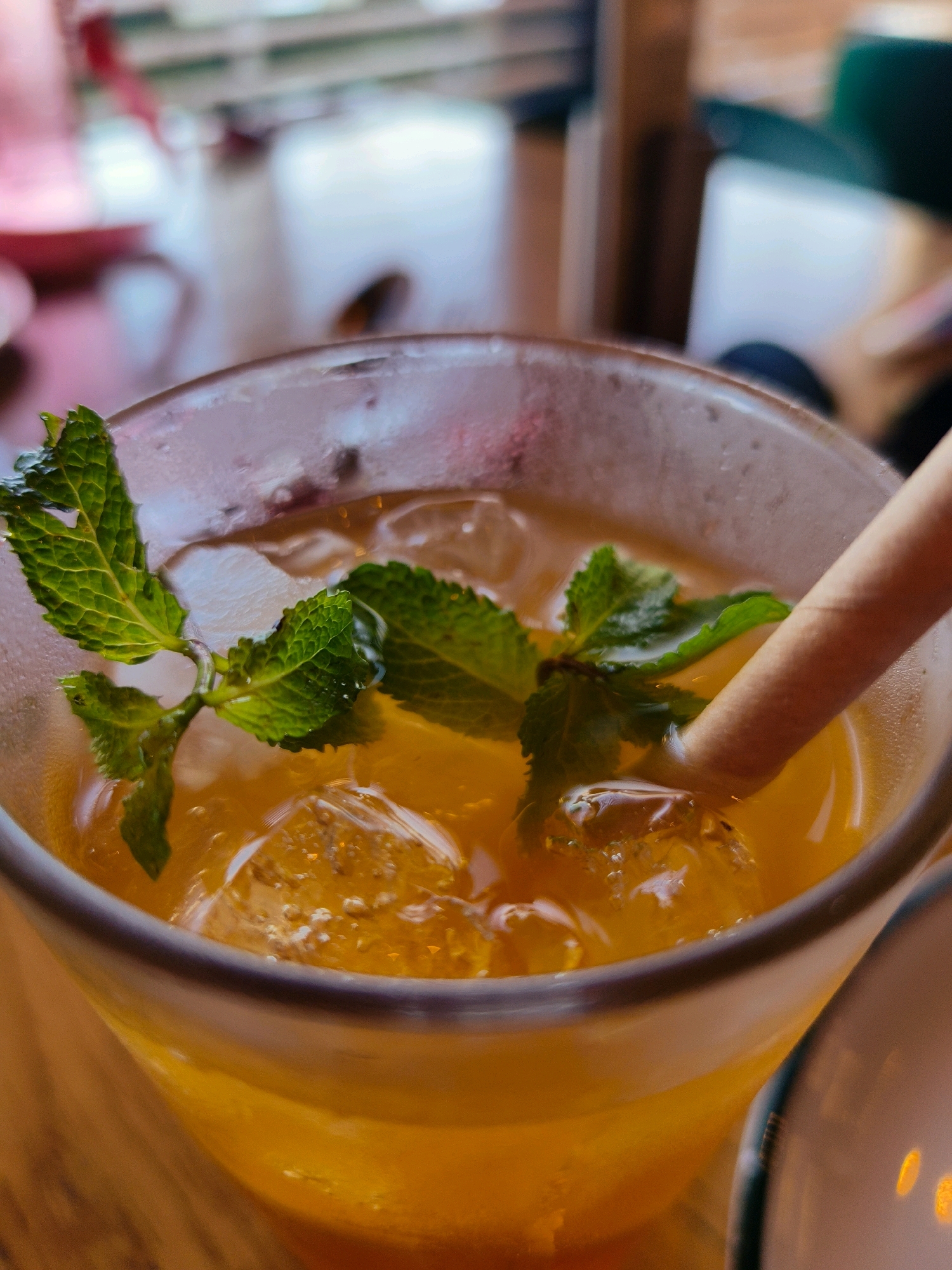 The image shows a close-up of a refreshing orange beverage in a clear glass, filled with ice cubes and garnished with fresh mint leaves. A light-colored straw is partially submerged in the drink. The background is blurred, suggesting an indoor setting, possibly a cafe or restaurant. The lighting is soft, highlighting the vibrant color of the drink and the texture of the mint. The image does not contain any identifiable landmarks or clues that would place it in Netivot, Israel, or any specific city. Therefore, the location in Netivot, Israel, cannot be determined from this image.