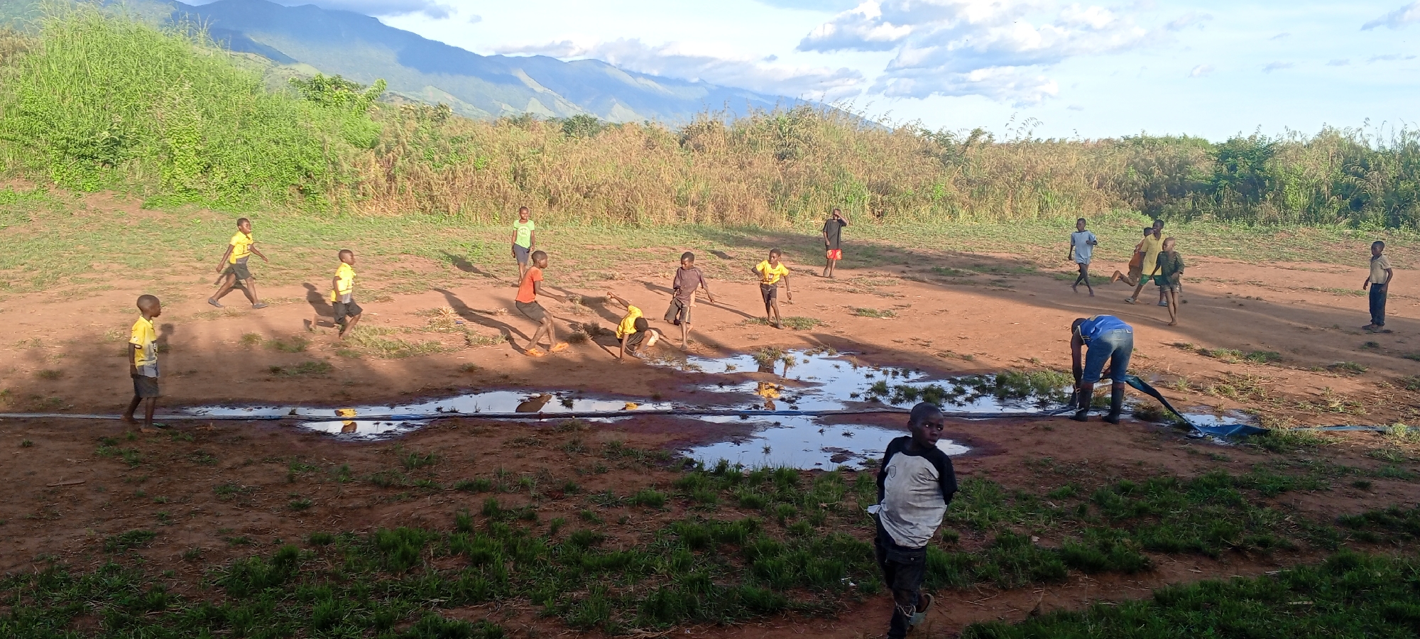 The image depicts a group of children in Ngalula, Democratic Republic of the Congo, on a dirt field with several puddles. Some children are playing in and around the puddles, while others stand or walk nearby. A man in a blue shirt and boots is handling a black hose near one of the puddles. The background includes grassy vegetation and mountains under a partly cloudy sky.