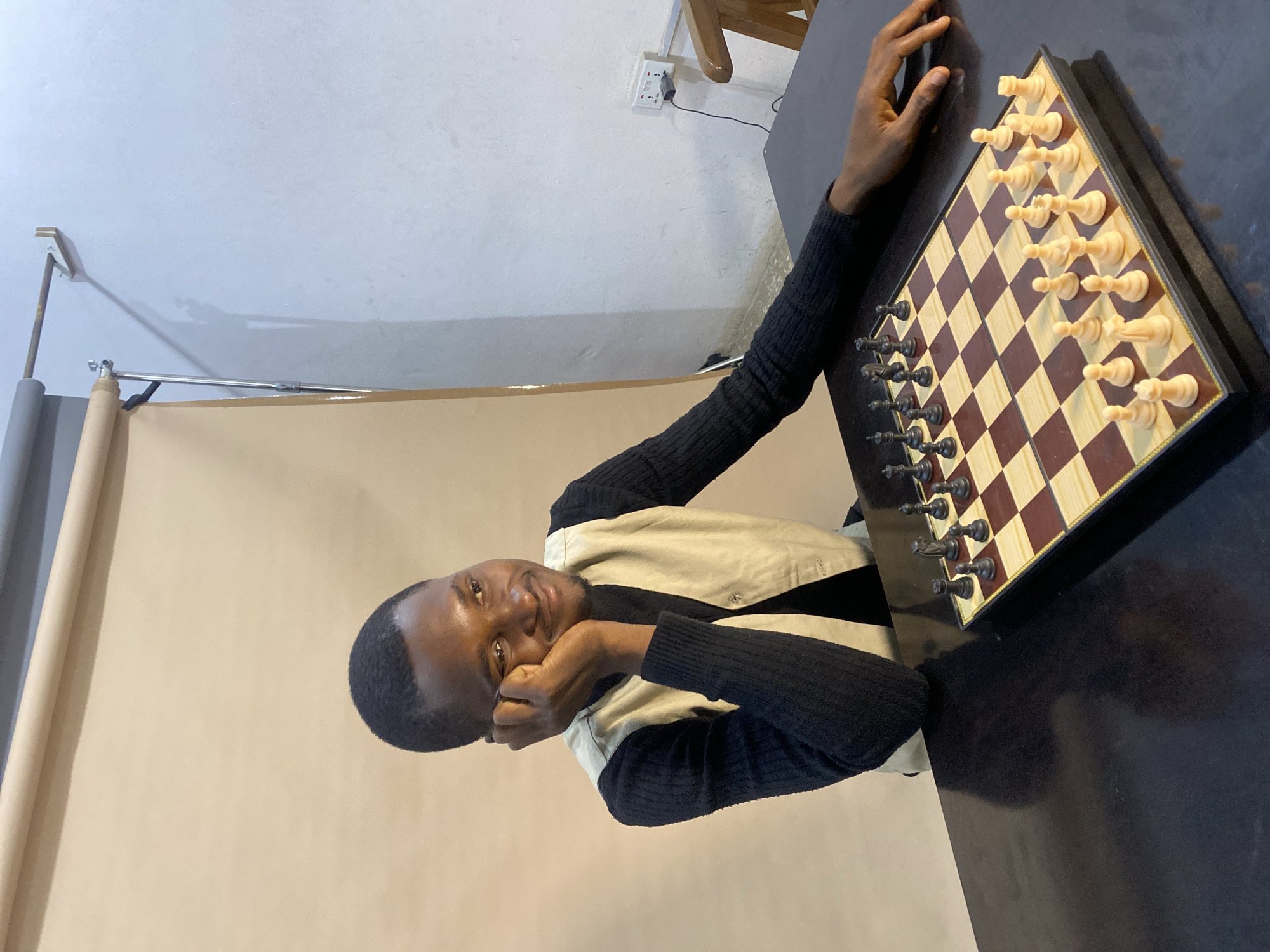 A young Black man is seated at a dark table with a chessboard. He is wearing a black turtleneck shirt and a light-colored vest. His right arm is resting on the table, and his right hand is supporting his chin as he looks to his left with a thoughtful expression. The chessboard is set up with pieces on both sides, and white pieces are in the foreground. The background consists of a beige backdrop and a white wall with a power outlet. The scene appears to be in an indoor studio or room. There is no specific indication of the location being in Gura Top, Nigeria.