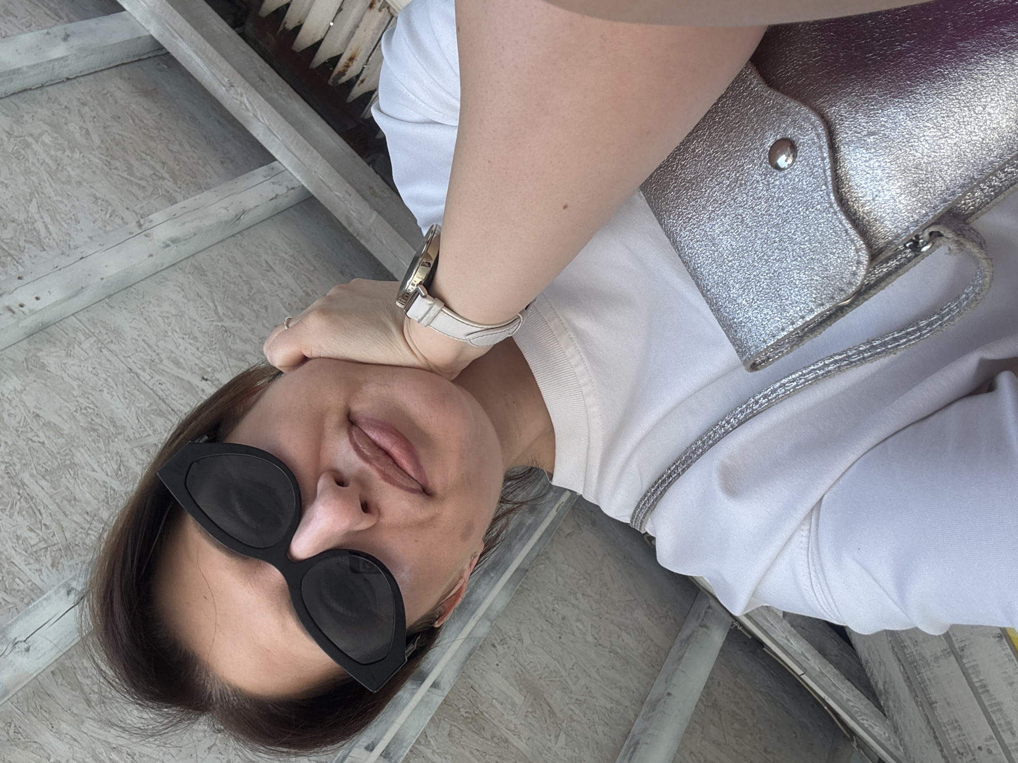 A woman in sunglasses and a white t-shirt is photographed from a low angle. She is touching her forehead with a ring on her finger. A silver purse hangs from her shoulder. The background includes gray wooden slats. The location is not discernable as being Sofia, Bulgaria.
