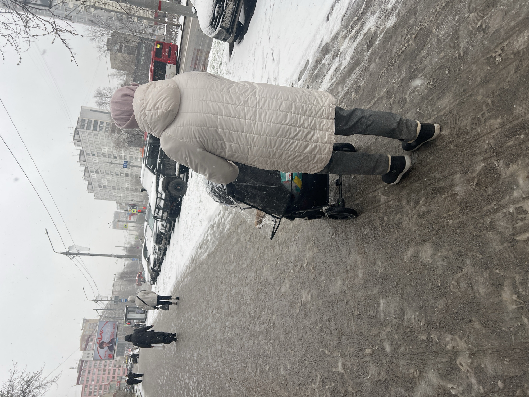 The image captures a winter street scene in Perm, Russia, during what appears to be daytime under overcast skies, with light snow falling. The ground is covered in a mixture of dirty snow, ice, and slush, showing numerous footprints and tire tracks.
The primary subject is a person, likely a woman, seen from behind, pushing a black baby stroller. She wears a light-colored (possibly white or very pale beige) quilted winter coat with a hood and dark grey pants, paired with black and white winter boots. The stroller has a transparent plastic cover.
In the background, a bustling city street unfolds. Parked cars, including an SUV with a visible Russian license plate "M382YK 159" (indicating Perm Krai), line the road. Further down, other pedestrians, dressed in dark winter clothing, are visible walking on the slushy sidewalk. A red bus can be partially seen in the distance on the street. The street is bordered by multi-story residential or commercial buildings, typical of post-Soviet architecture, with some bare trees dotting the landscape. Overhead, power lines crisscross the view.
A large billboard is visible further down the street on the right, displaying text that includes "АЛЫЕ ПАРУСА" (Alye Parusa, meaning 'Scarlet Sails') and an image of a man. The overall atmosphere is that of a cold, ordinary winter day in an urban environment.