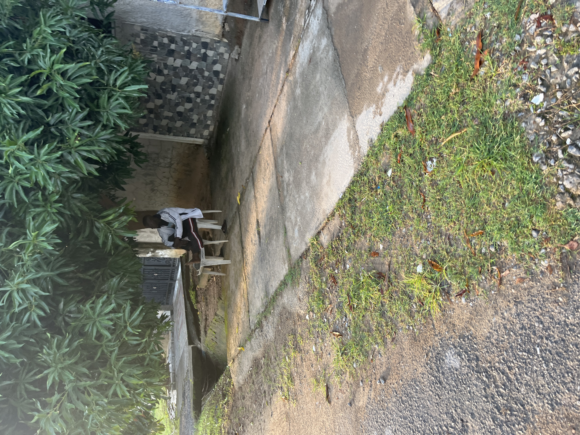 A person is sitting on a white plastic chair under the shade of a large tree. The person is wearing a white jacket with black trim and black pants. A low concrete wall with a textured, mosaic-like pattern is visible behind the person. In front of the person, there is a concrete walkway and a shallow, overgrown drainage ditch. The ground is a mixture of dirt, grass, and small stones. The scene appears to be in an outdoor area, possibly a residential street or a public space, in Abuja, Nigeria, indicated by the presence of a prominent tree and the architectural style.