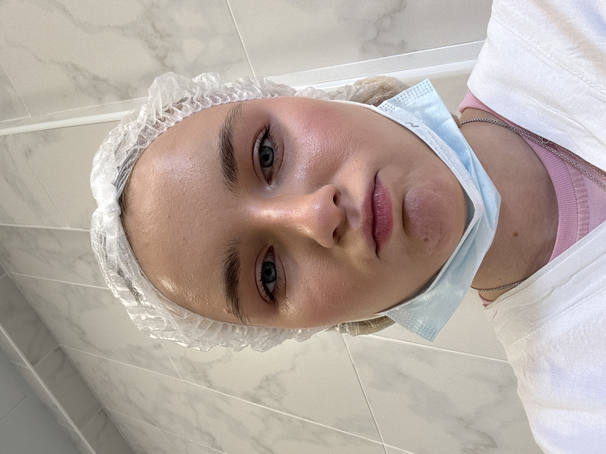 A young woman is seen from an upward, slightly oblique angle. She wears a white, disposable hairnet and a light blue surgical mask pulled down below her chin. Her clothing includes a white garment and a pink shirt, along with a silver necklace. The background consists of light-colored marble-patterned tiles. There is no information to indicate that the image was taken in Saint Petersburg, Russia.