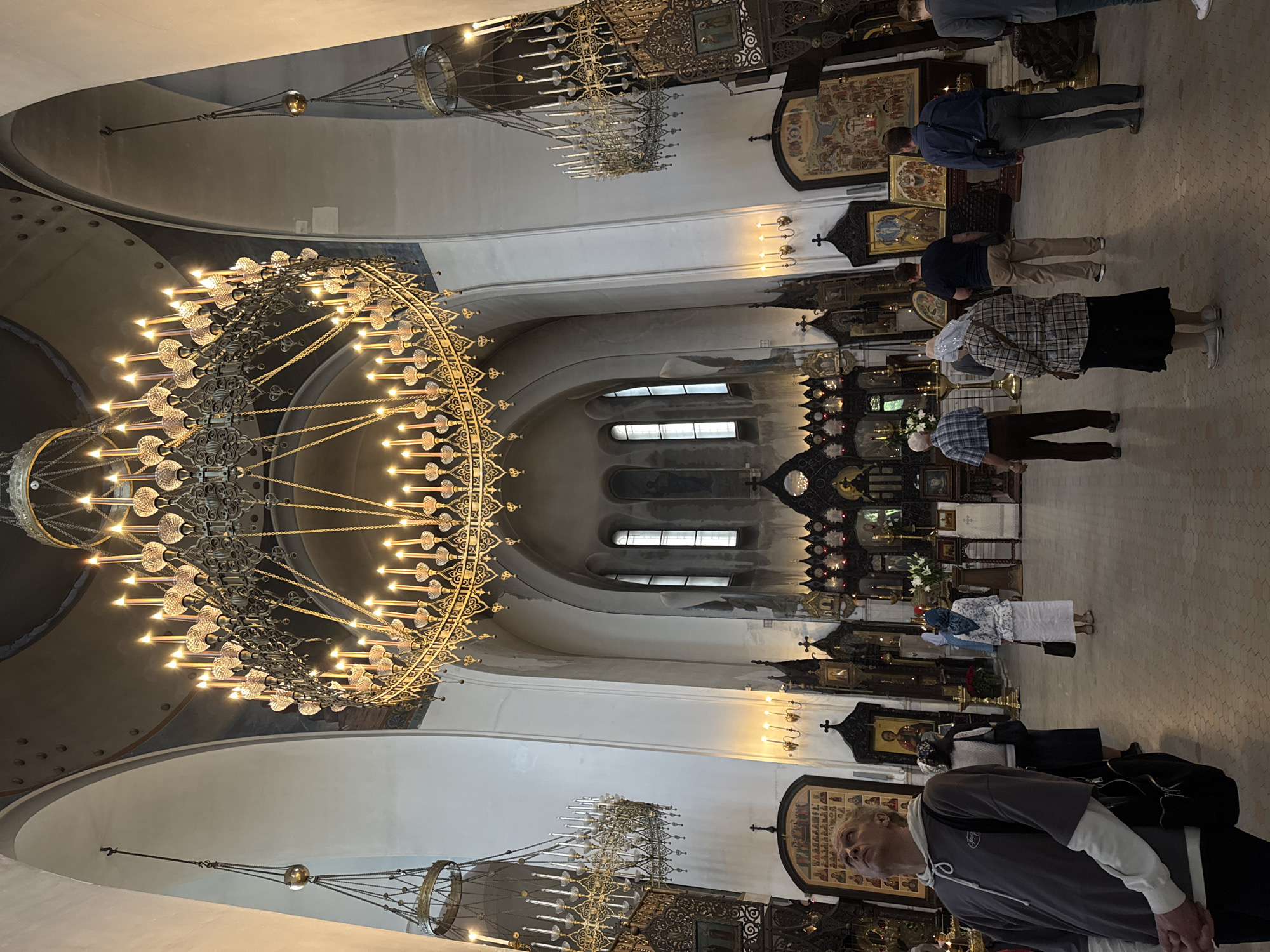 The image captures the interior of an Orthodox church, likely in Moscow, Russia, given the architectural style and the presence of religious icons and artifacts. Dominating the scene is a large, ornate chandelier with numerous lit bulbs, suspended from the high ceiling. The church features arched ceilings and a traditional iconostasis – a wall of icons separating the nave from the sanctuary. Several people are present in the church, some of whom appear to be praying or observing religious rituals. The lighting is a mix of natural light entering through arched windows and the warm glow from the chandelier. The overall atmosphere is reverent and contemplative.