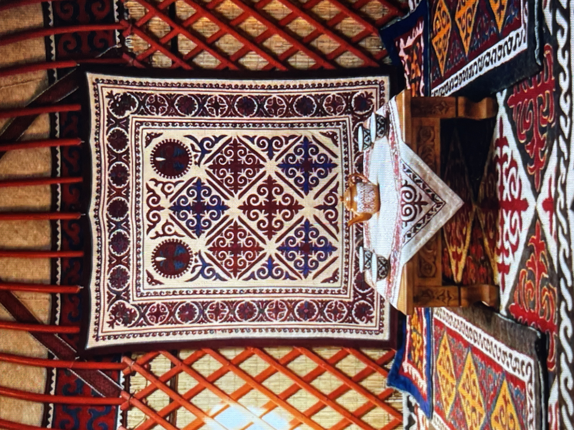 The image depicts the interior of a yurt, a traditional dwelling, likely located in or near Bishkek City, Kyrgyzstan. The scene is richly decorated with textiles. Dominating the background is a large, ornately patterned tapestry hanging on the wall, featuring geometric motifs in burgundy, cream, and blue. The yurt's lattice walls, made of wood and painted red, are visible on the sides, with red poles extending upwards. In the foreground, a low wooden table is set with a teapot and two small cups, all arranged on a white tablecloth with a decorative border. The floor is covered with multiple colorful carpets, showcasing intricate patterns in red, yellow, blue, and black. The overall impression is one of traditional craftsmanship and cultural heritage. There are no people visible in the image.