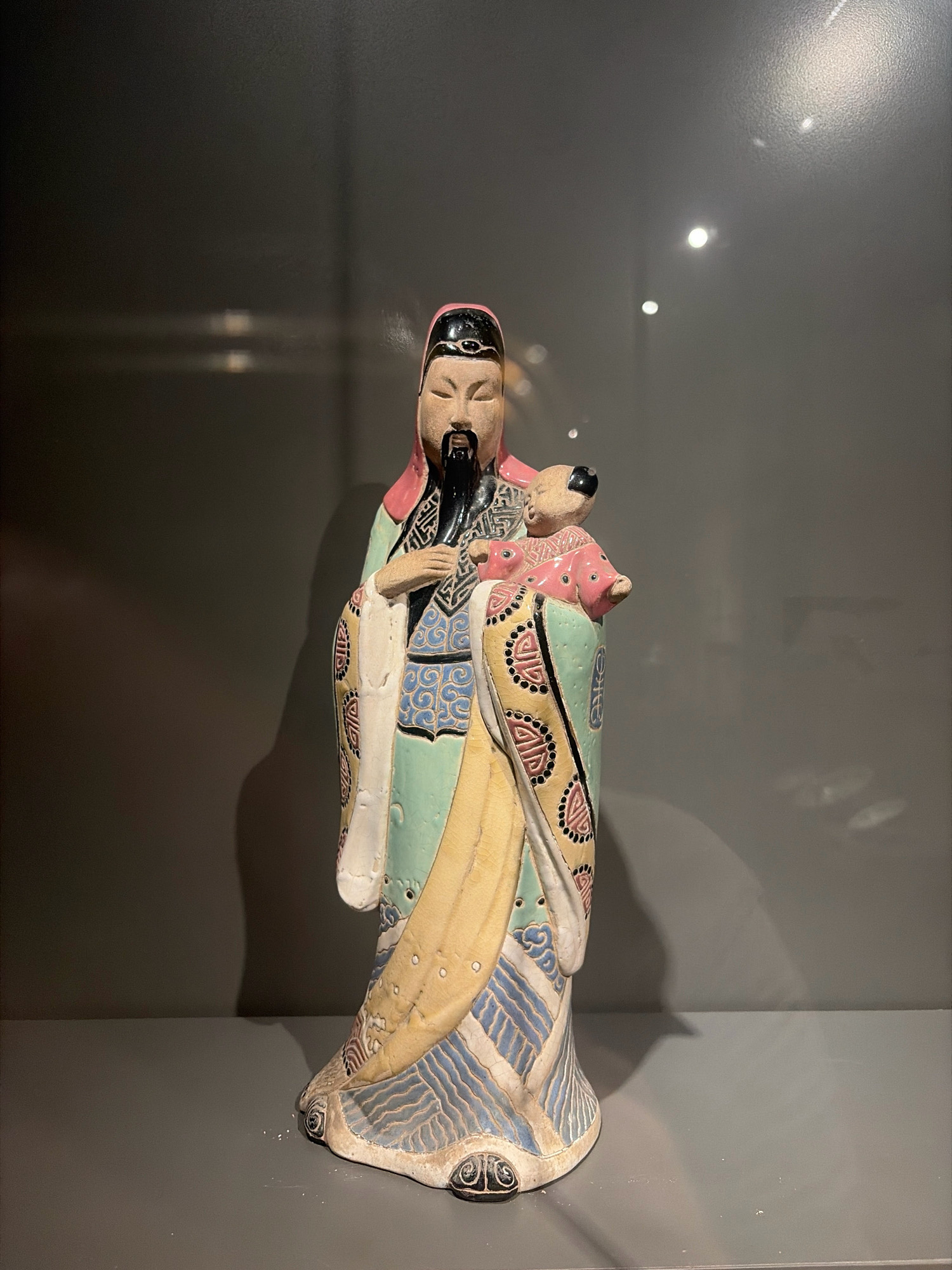 The image shows a full-body, front-facing ceramic figurine of an elderly East Asian man with a long black beard, wearing a pink cap, a light green robe with colorful patterns, and a white underskirt. He is holding a pink baby with a black head in his arms. The figurine is standing on a grey surface, with a grey wall in the background. The lighting creates reflections and shadows on the wall. The figurine appears to be displayed in a museum or exhibition. The image is taken from a medium distance. This scene is not located in the city of Đà Nẵng, Vietnam.