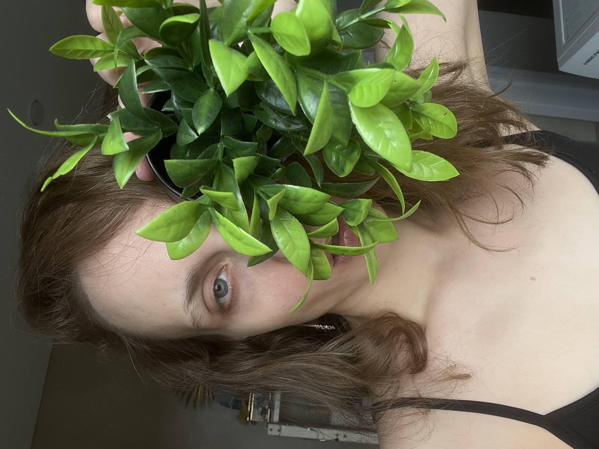 A woman with fair skin and brown hair holds a potted green plant in front of her face. Her left eye is visible through the leaves. She is wearing a black tank top. The background appears to be an indoor space, possibly a home or office, in a city and country that are unknown.