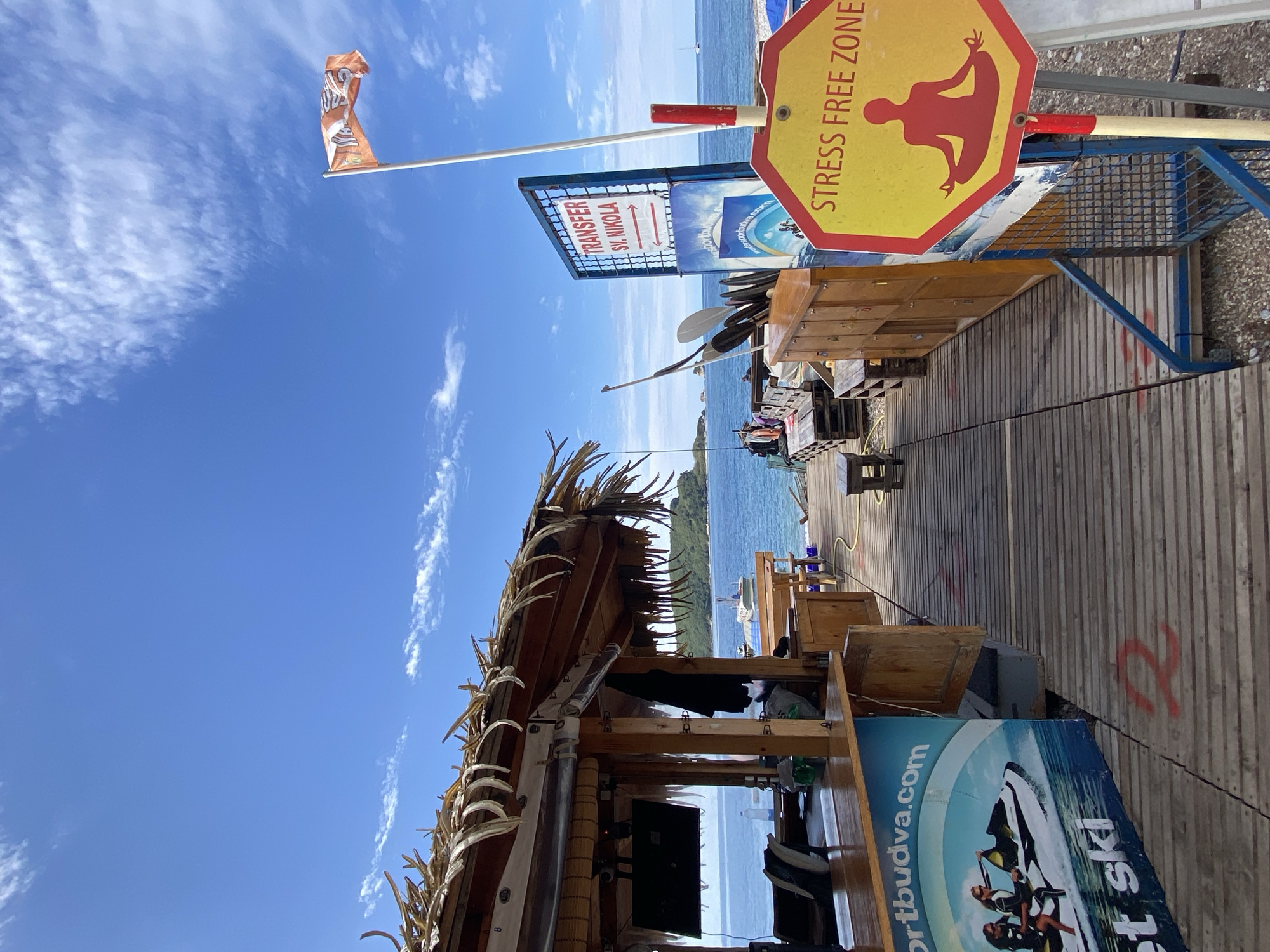 The image shows a beach bar/recreation area in Budva, Montenegro. The foreground has a wooden structure with a thatched roof, likely a bar or concession stand. The roof is adorned with decorative elements. A sign featuring jet skis and the website "sportbudva.com" is partially visible. A "TRANSFER ST. NIKOLA" sign indicates a transportation service, possibly to a nearby island. A yellow, octagonal "STRESS FREE ZONE" sign with a silhouette of a person in a meditative pose is present. In the background, the Adriatic Sea is visible, along with a distant shoreline or island. Several people can be seen enjoying the beach and water activities. The sky is blue with scattered clouds. A flag is also visible in the sky. The overall impression is of a sunny, relaxed beachside environment catering to tourists.